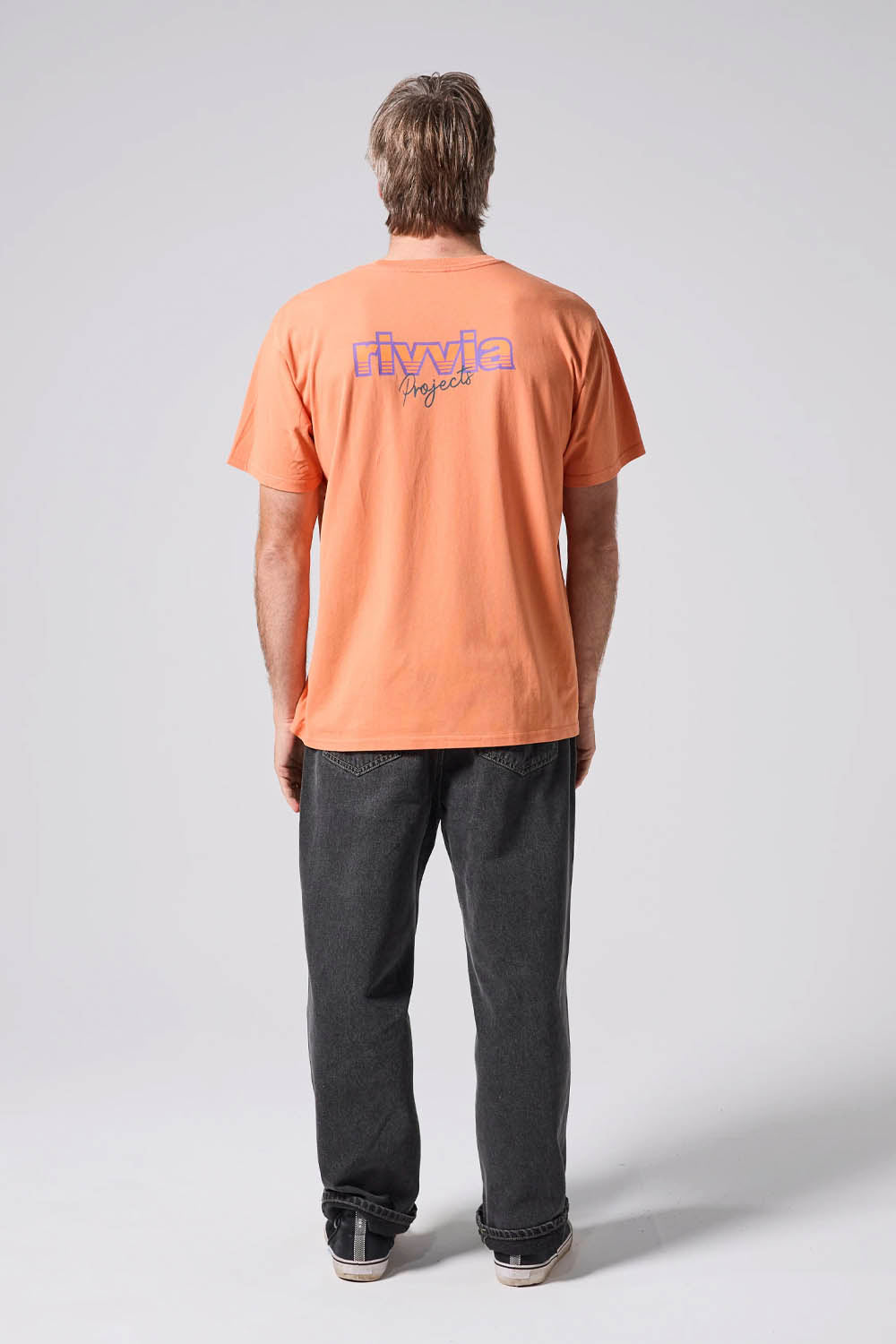 Person wearing a peach t-shirt with a logo on the back, standing against a white background