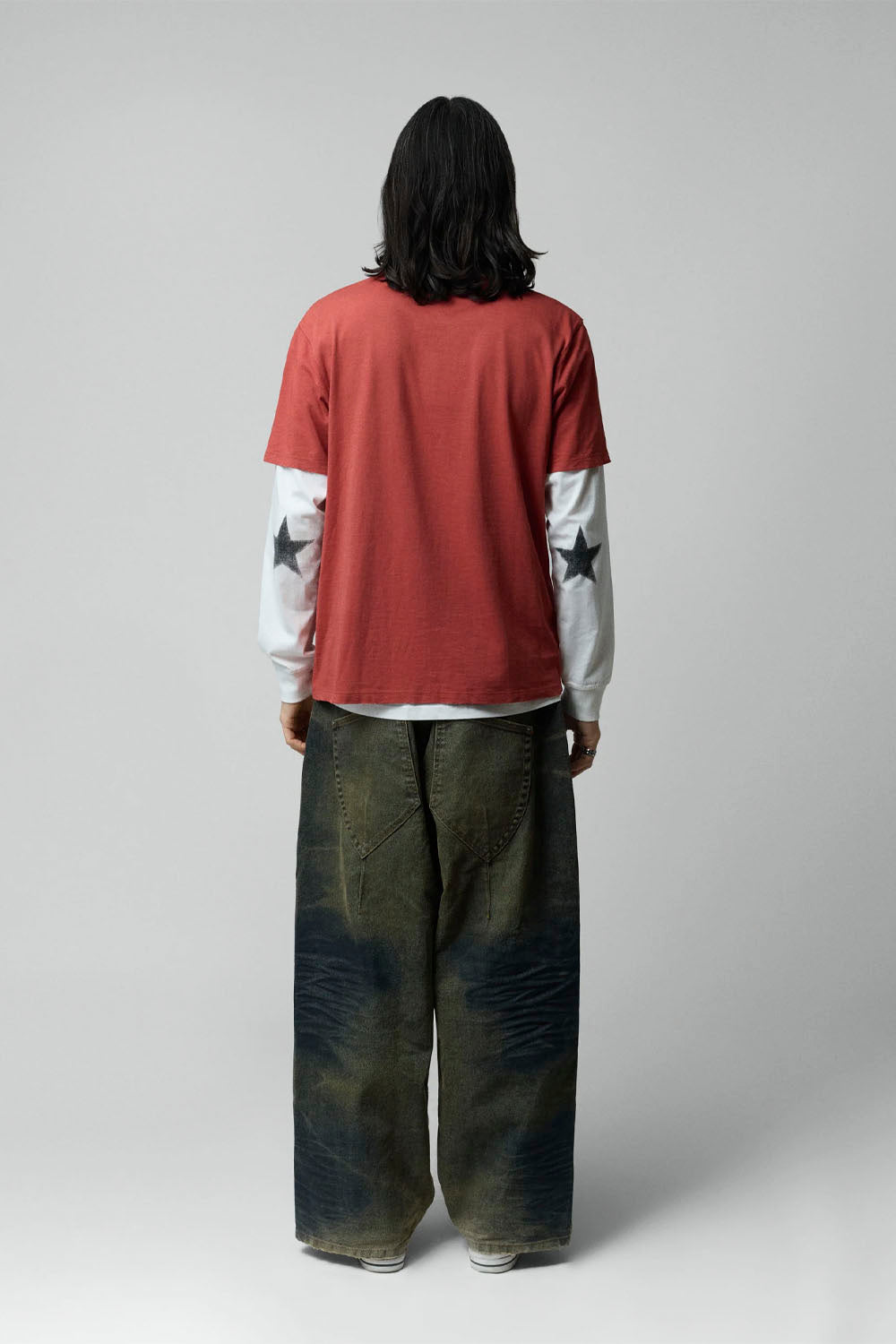 A model viewed from the back wearing a red short-sleeve t-shirt layered over a white long-sleeve shirt with black star graphics on the sleeves, paired with wide-leg dark green and blue tie-dye jeans, against a light gray studio background.