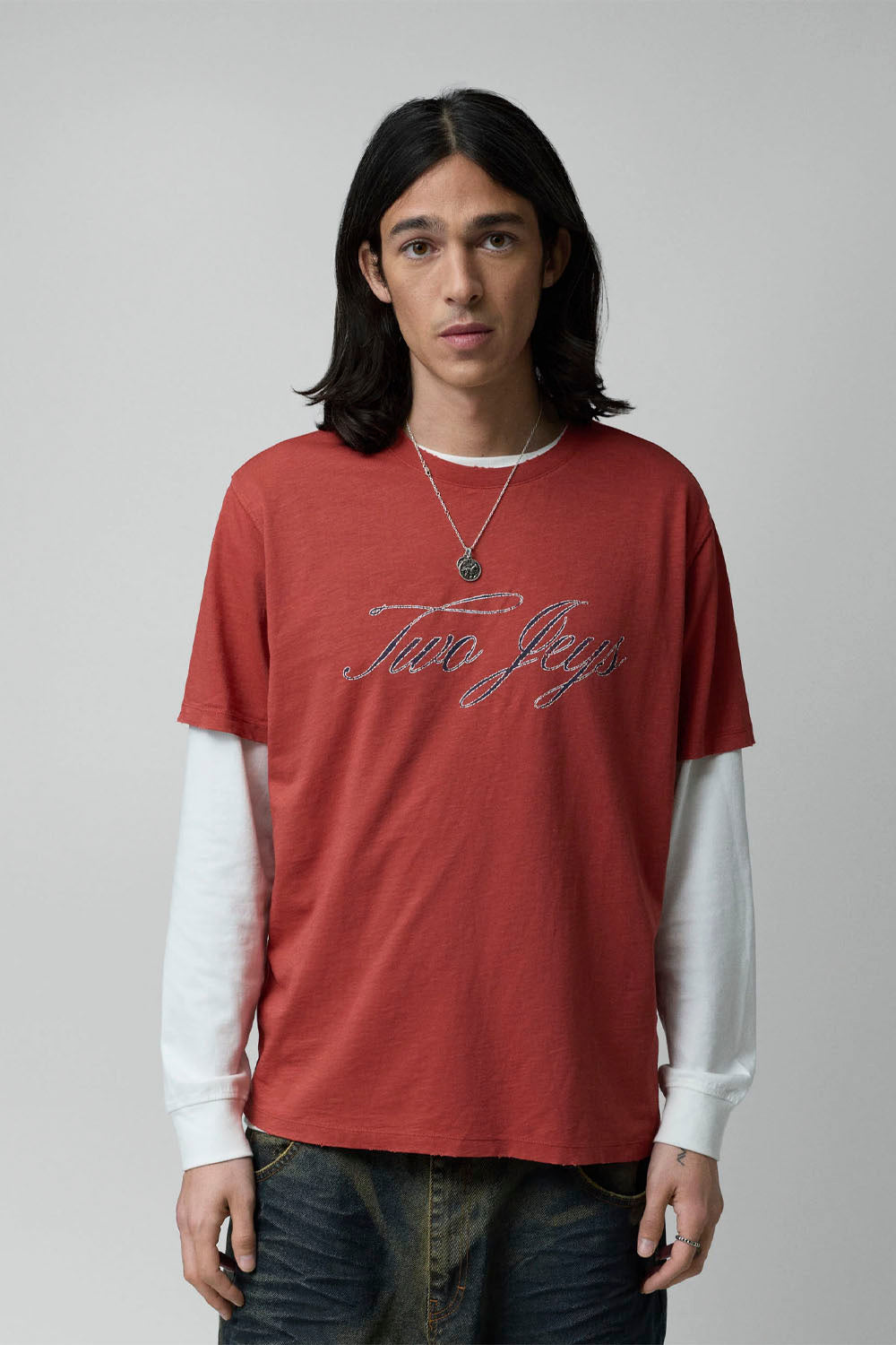 A model wearing a rust-red short-sleeve t-shirt with 'Two Jezz' written across the chest in metallic script, layered over a white long-sleeve shirt and dark blue jeans, against a solid gray background.