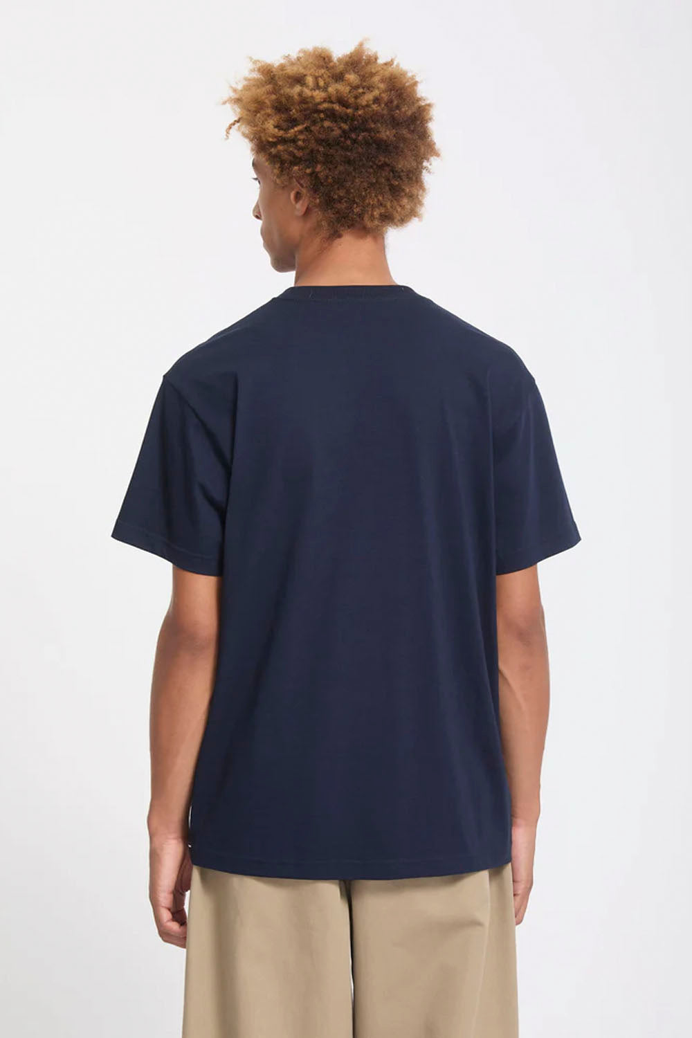 A man with curly light brown hair is shown from the back wearing a navy blue short-sleeve t-shirt and beige trousers against a plain white background.
