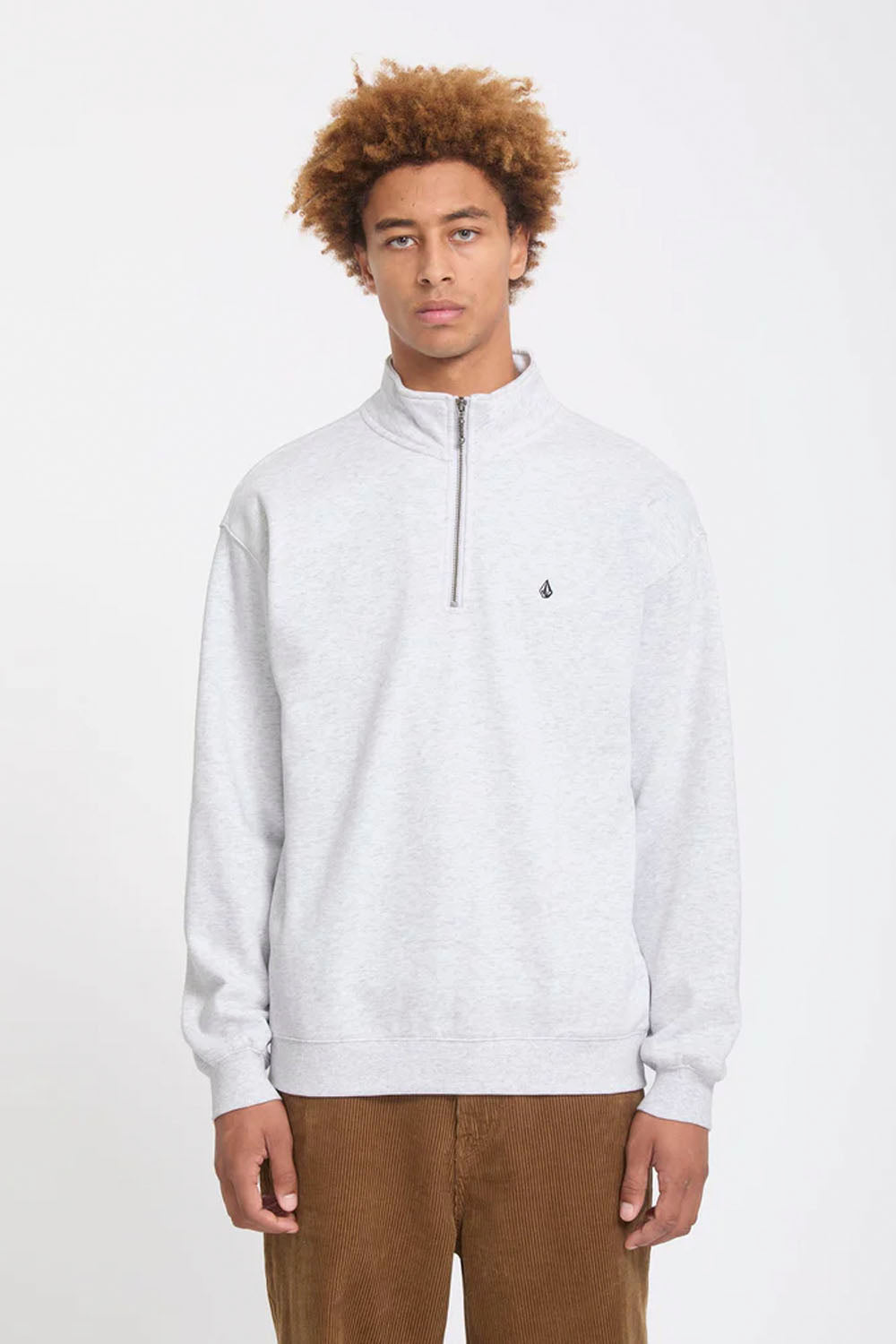 A man wearing a light gray heathered quarter-zip sweatshirt with a small black Volcom logo on the chest, paired with brown pants, against a plain white studio background.