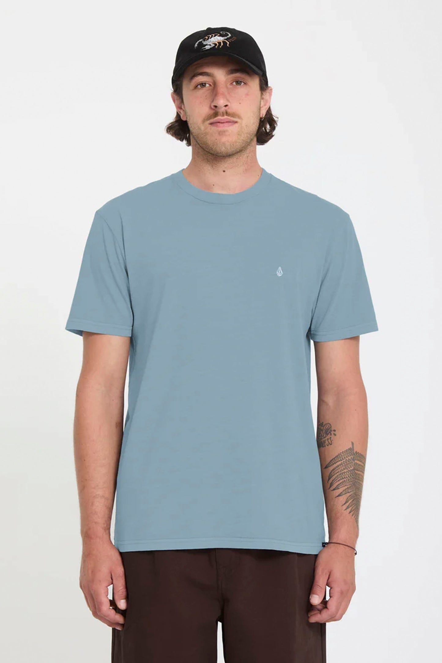 Man wearing a light blue Volcom Solid Stone t-shirt and black cap on a white background
