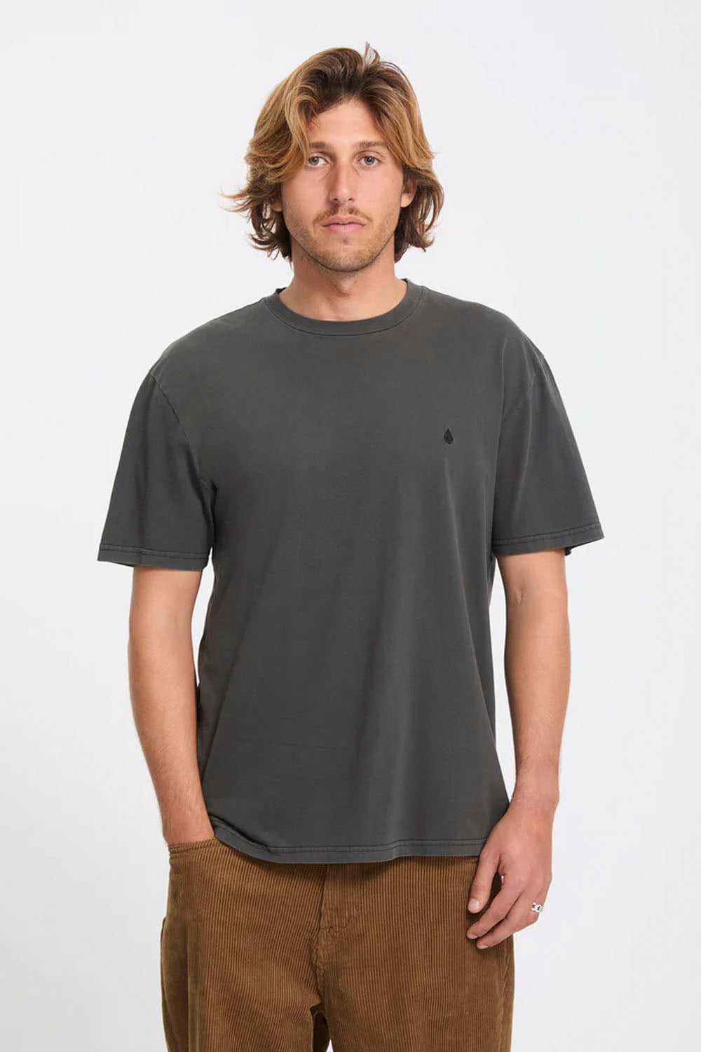 A man wearing a dark charcoal gray short-sleeve crewneck t-shirt with a small black embroidered polo player logo on the left chest, paired with brown pants, against a plain light gray background.