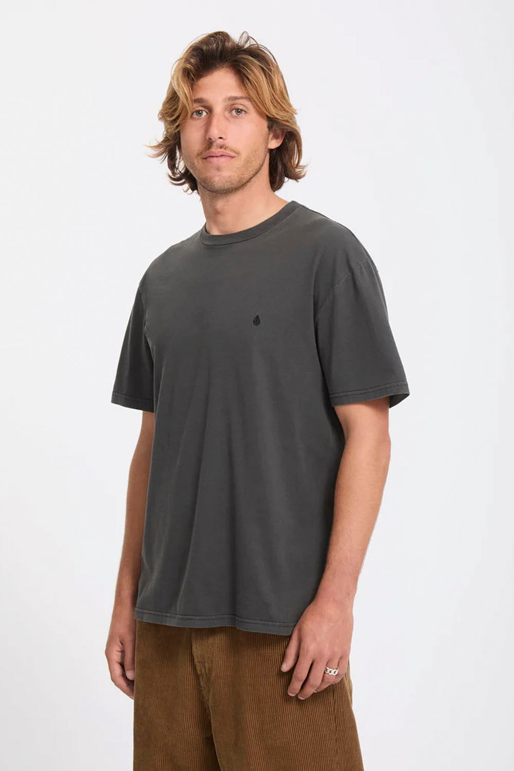 A man wearing a dark charcoal gray short-sleeve crewneck t-shirt with a small black embroidered logo on the left chest, paired with brown corduroy pants, photographed against a plain white studio background.