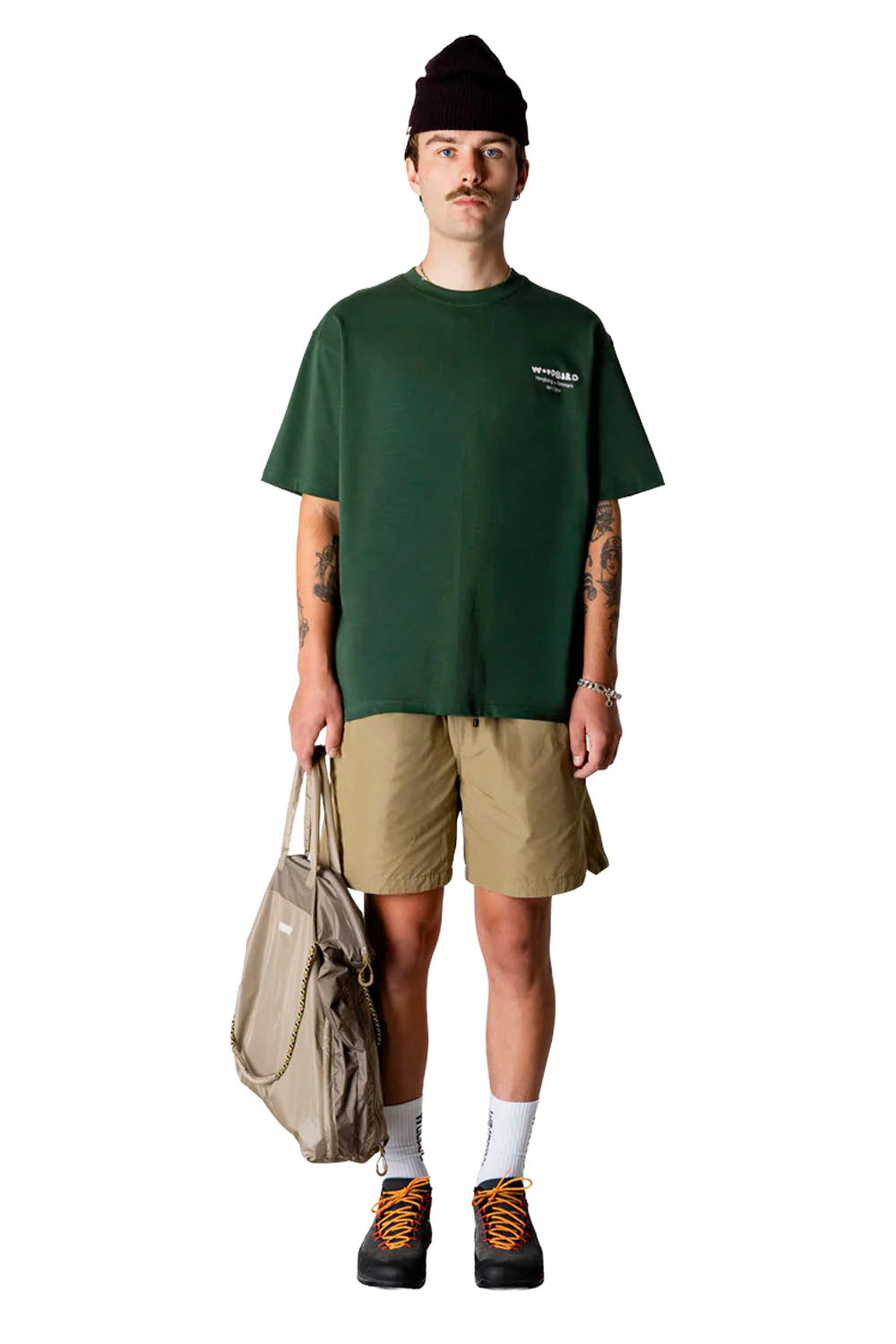 Man wearing a green t-shirt, beige shorts, and a beanie, holding a beige bag on a white background