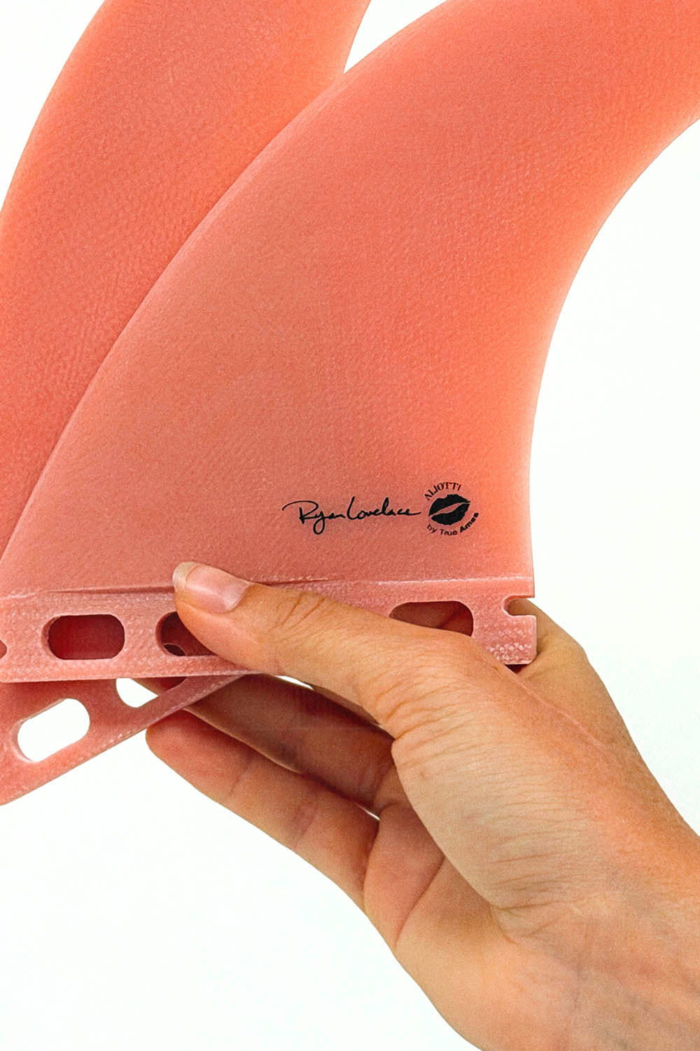 A close-up of a person's hand holding a set of pink surfboard fins against a white background, showing the 'Ryan Lambini' brand signature printed on the fin.