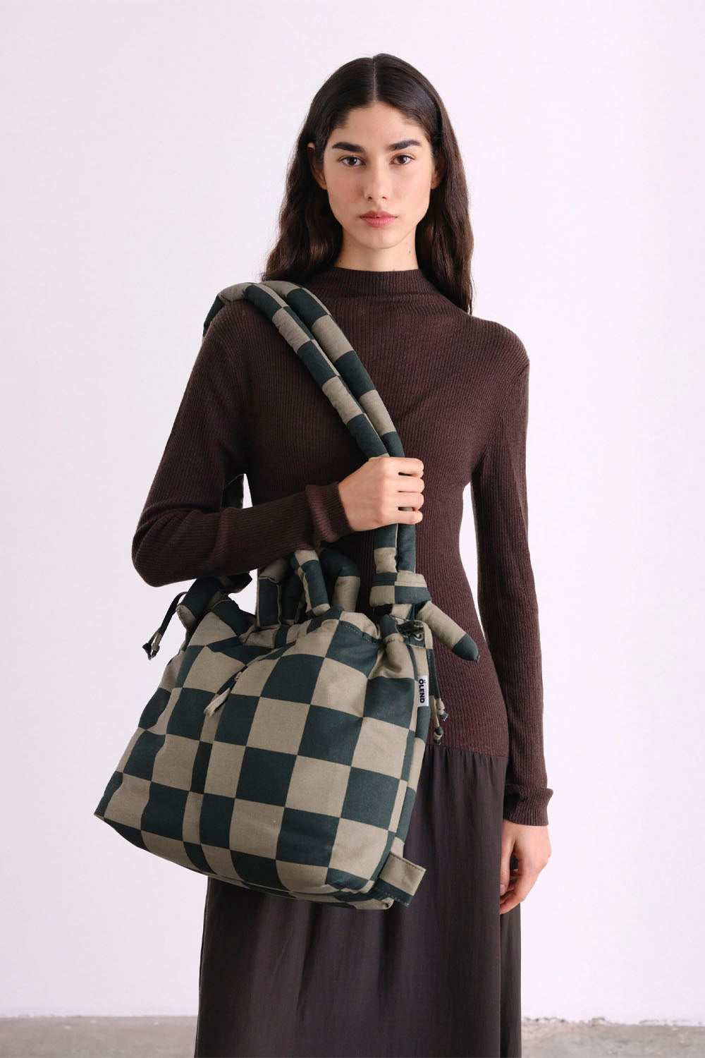 A model wearing a dark brown mock-neck sweater and dark skirt holds a large checkered canvas shoulder bag in green and beige. The background is a clean, off-white studio setting.