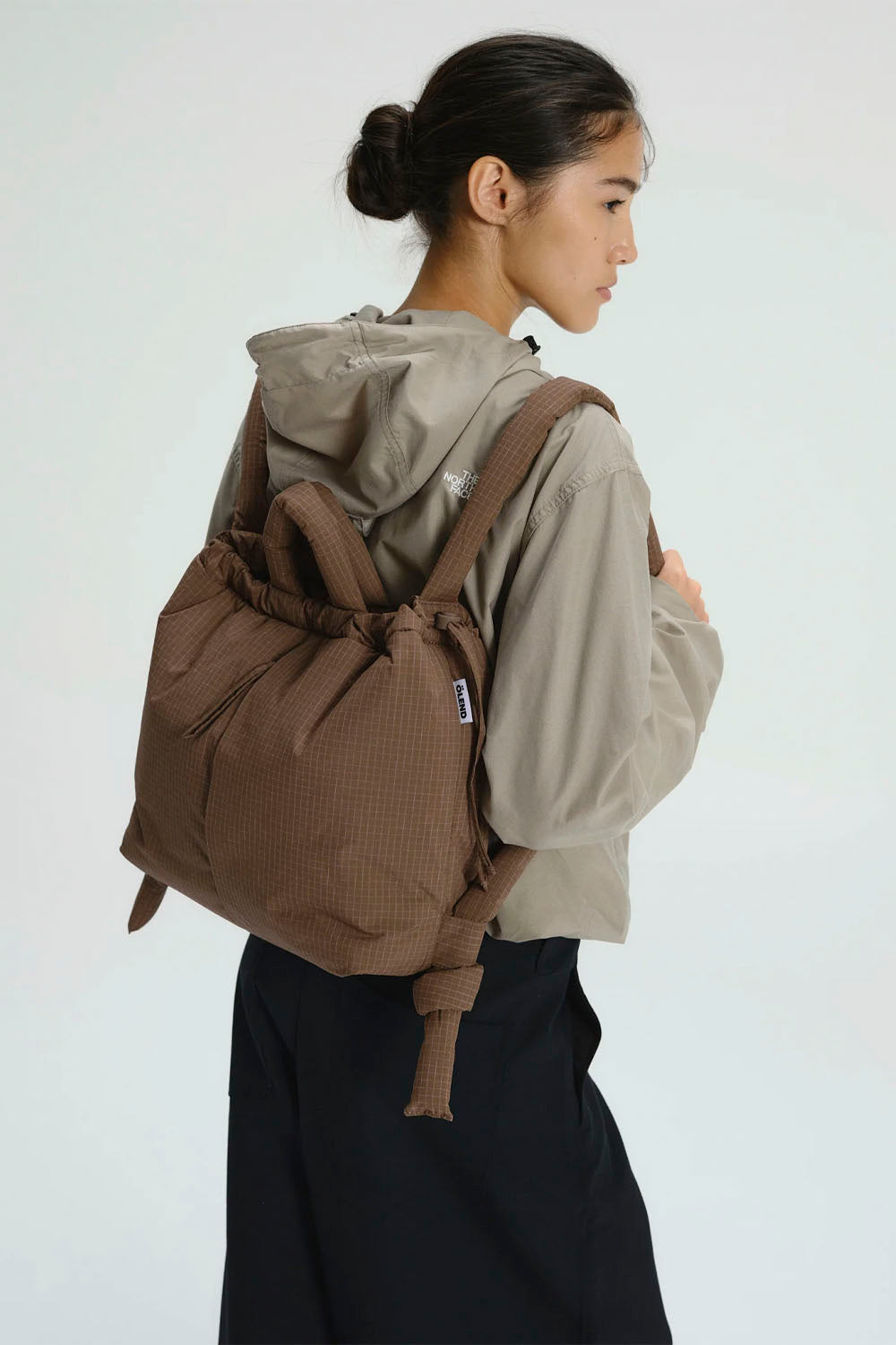 A woman shown from the back wearing a brown textured drawstring backpack over a beige hooded jacket and black skirt, against a plain light gray studio background.