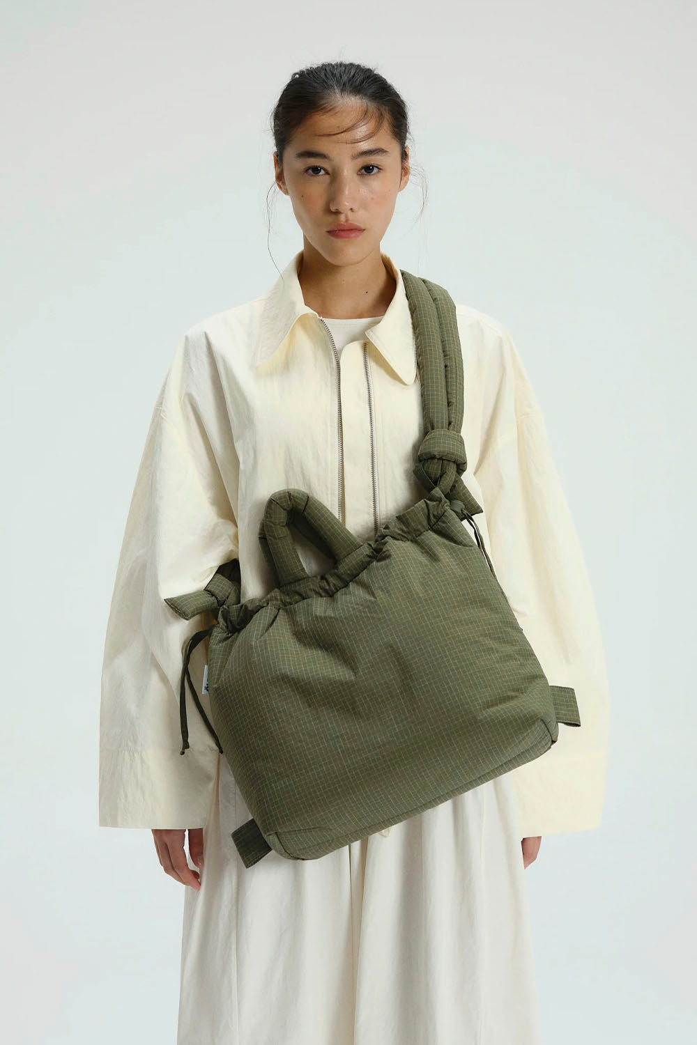 A model wearing a cream-colored shirt and white skirt, carrying an olive green textured drawstring bucket bag over her shoulder, set against a plain light gray background.