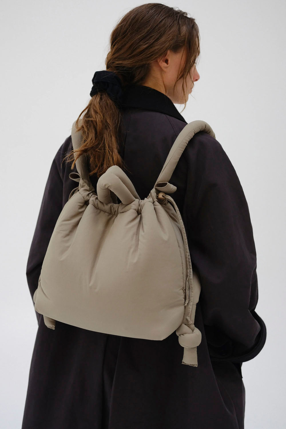 A woman shown from behind wearing a light beige puffer backpack with knotted shoulder straps over a dark coat, against a plain light gray background.