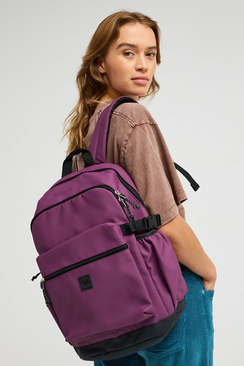 A woman wearing a brown t-shirt and blue corduroy pants carries a purple and black backpack on her shoulder against a light gray studio background.
