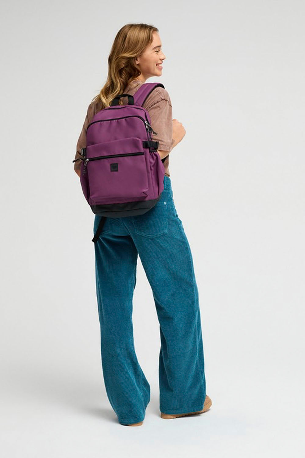 A woman wearing a purple backpack, brown top, and teal corduroy pants, photographed from the back against a light gray studio background.