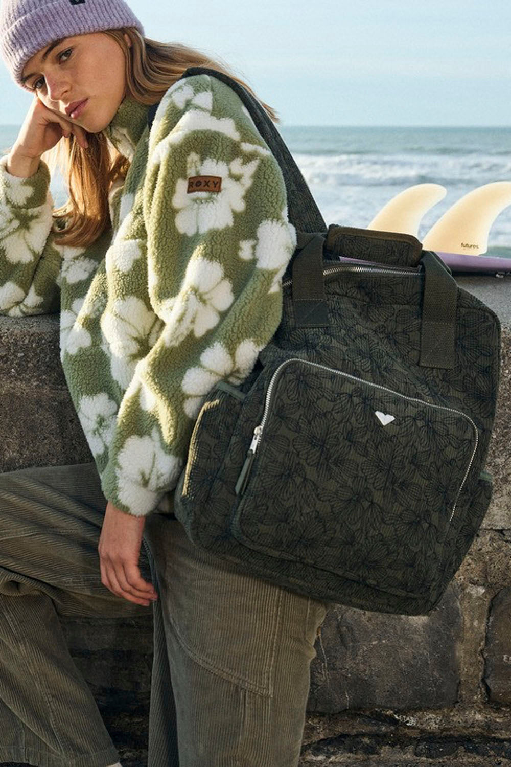 A person standing on a stone pier by the ocean, wearing a dark olive backpack, a green floral jacket, and a purple beanie, with one surfboards in the background.