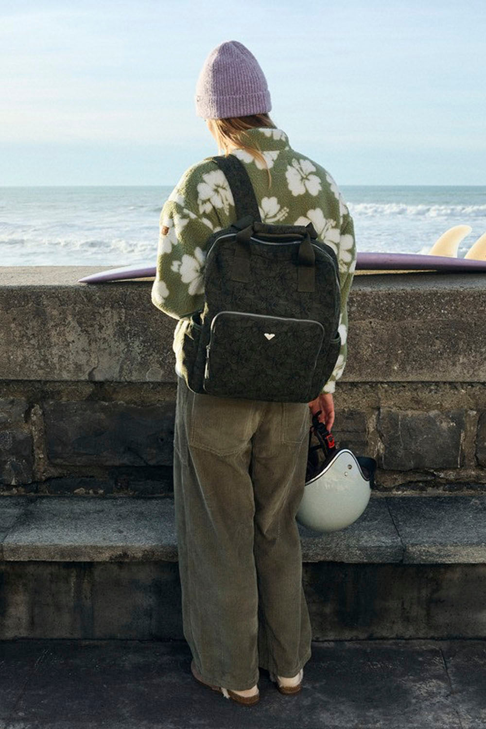 A person standing on a stone pier by the ocean, viewed from behind, wearing a dark olive backpack, a green floral jacket, and a pink beanie, with a white helmet in one hand and surfboards in the background.
