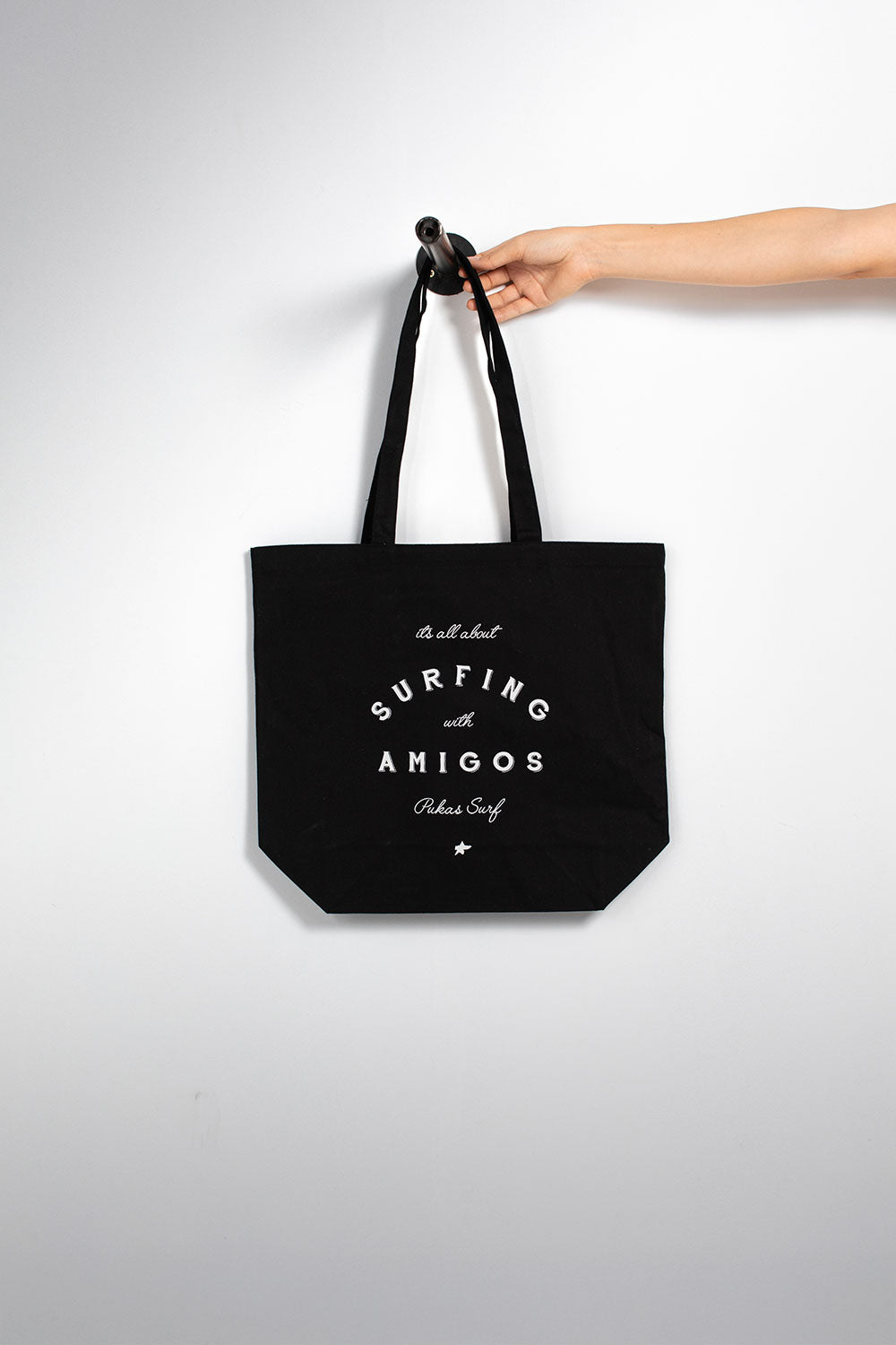 black canvas tote bag. The bag features white printed text that reads ' surfing with AMIGOS' and 'Pukas Surf'.
