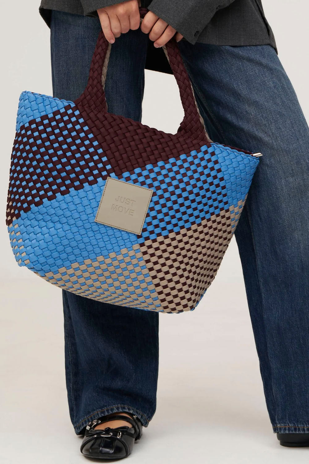 A person wearing blue jeans and black shoes holds a large woven tote bag with geometric color blocks in blue, dark brown, beige, and gray. The bag has a square metal brand plaque on the front that reads 'JUST MOVE'.