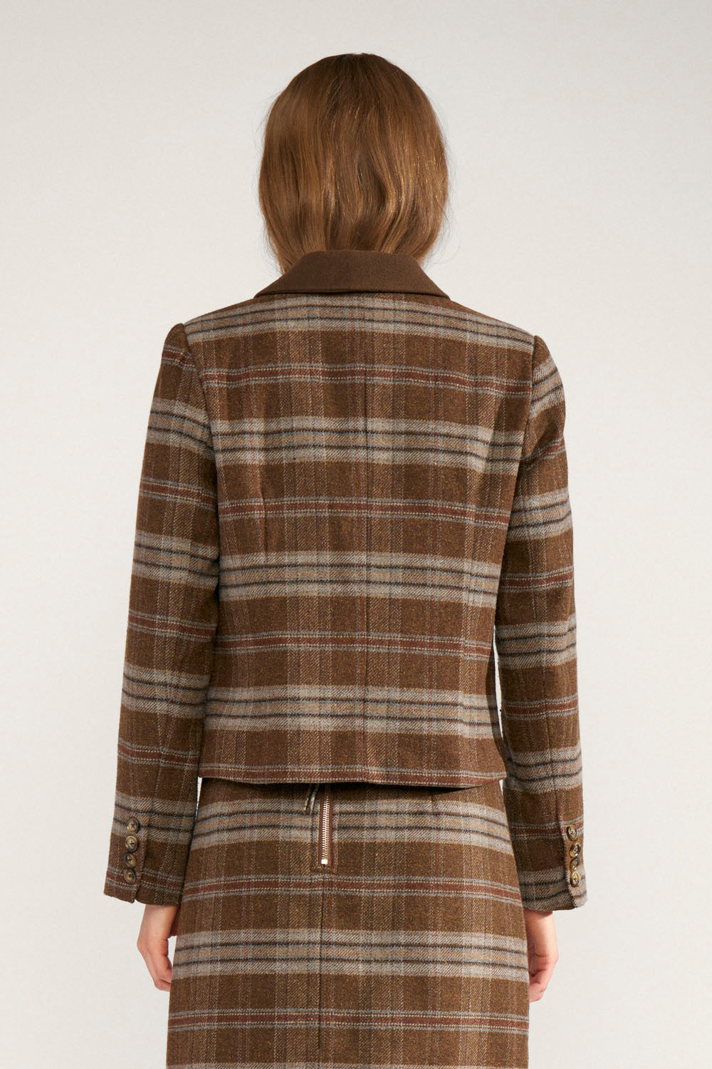 A model viewed from the back wearing a brown plaid cropped jacket and matching skirt against a plain light background.
