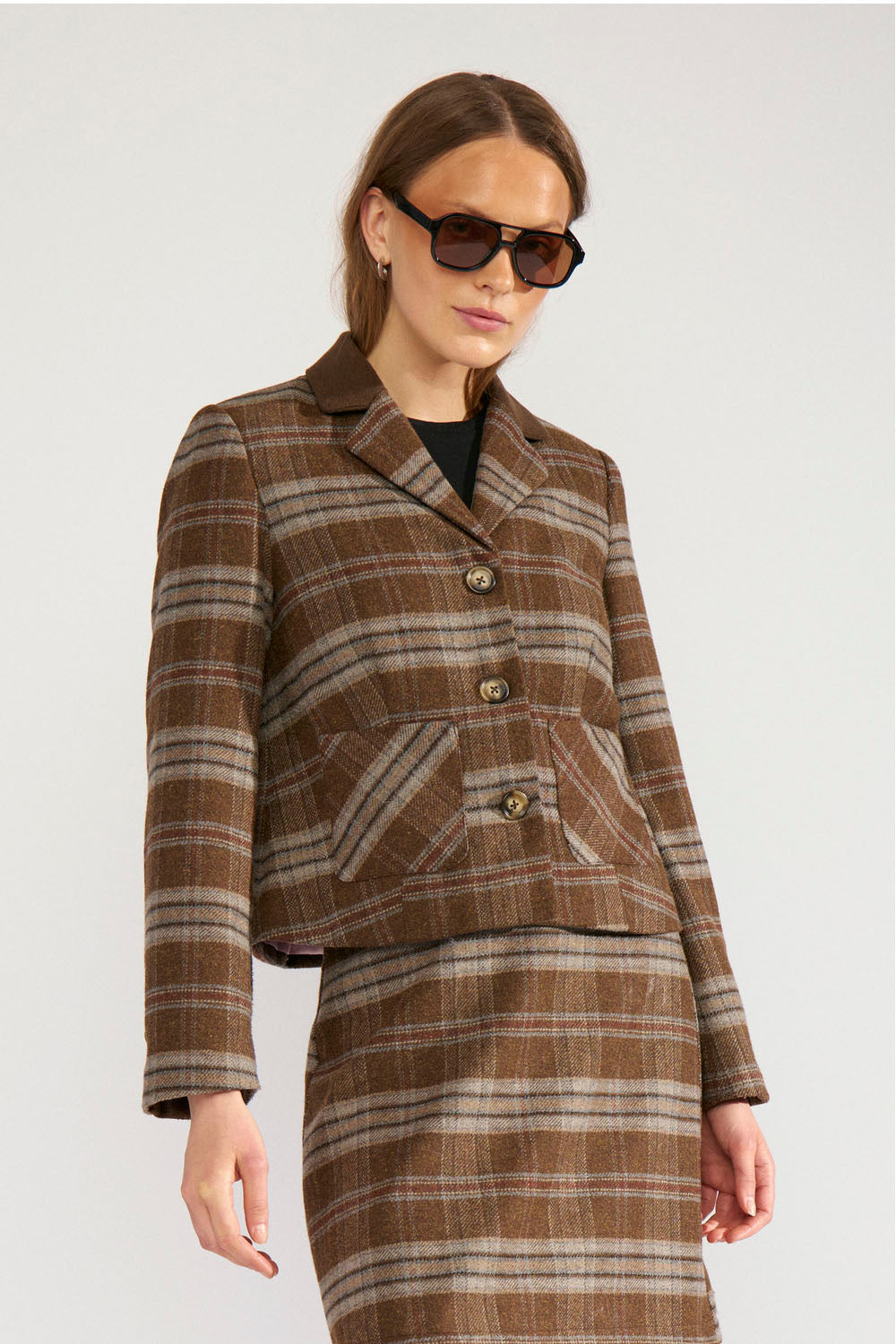 A woman modeling a brown and beige plaid tweed jacket and matching skirt set, styled with dark sunglasses against a light gray studio background.