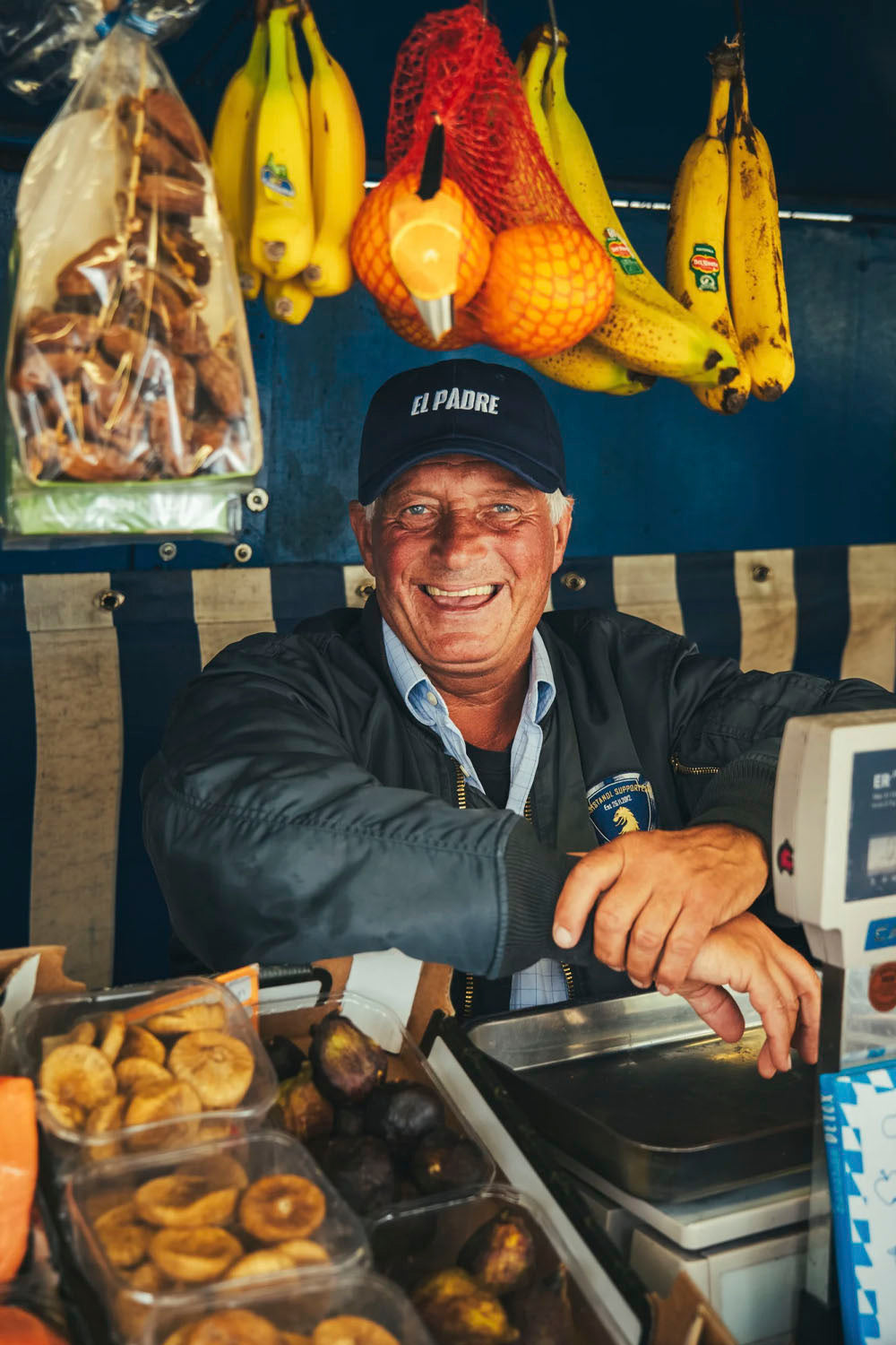 A smiling man wearing a black cap that reads 'EL PADRE' sits behind a fruit and bread stand, with bananas, oranges, and trays of pastries visible in the scene.