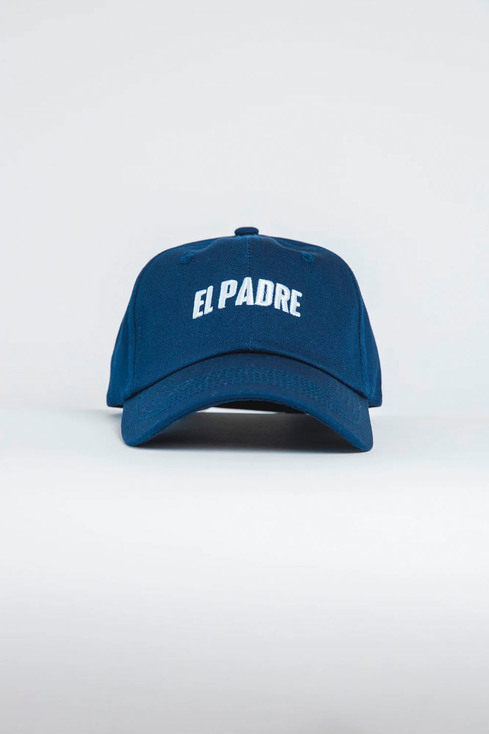 A blue baseball cap with 'EL PADRE' embroidered in white arched letters on the front, photographed against a light gray studio background.