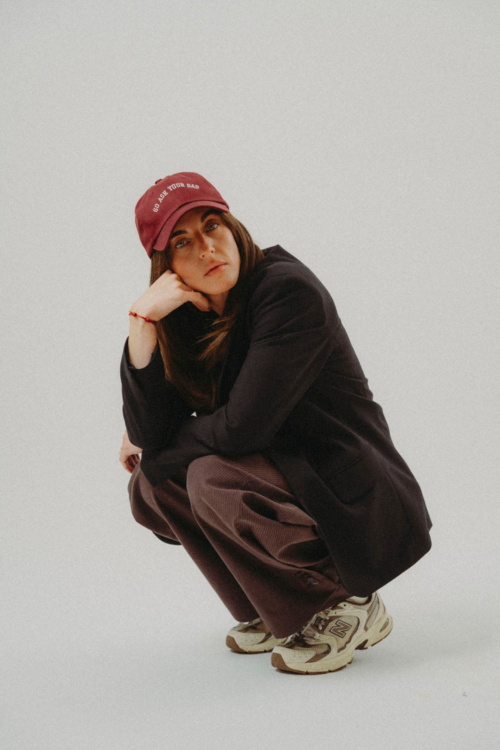 A model crouching in an urban pose, wearing a maroon baseball cap with white text, a dark brown long-sleeve top, matching dark brown wide-leg pants, and beige and brown sneakers, against a solid light gray background.