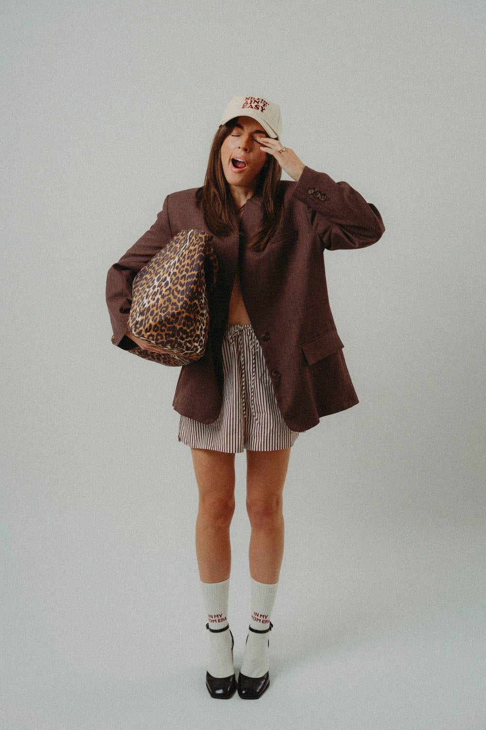 A woman posing in a studio setting wearing an oversized dark brown blazer, a beige and white striped pleated mini skirt, white socks, and black shoes. She is holding a leopard print shoulder bag and wearing a beige baseball cap with red lettering.