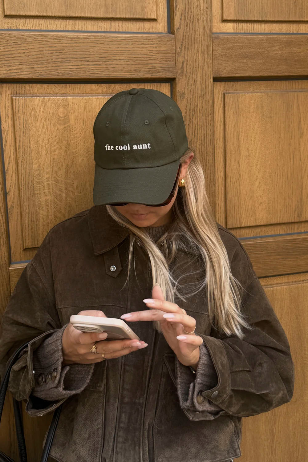 A woman wearing an olive green baseball cap with the text 'the cool aunt' embroidered in white, paired with a brown corduroy jacket, looking down at a phone in front of a wooden door.