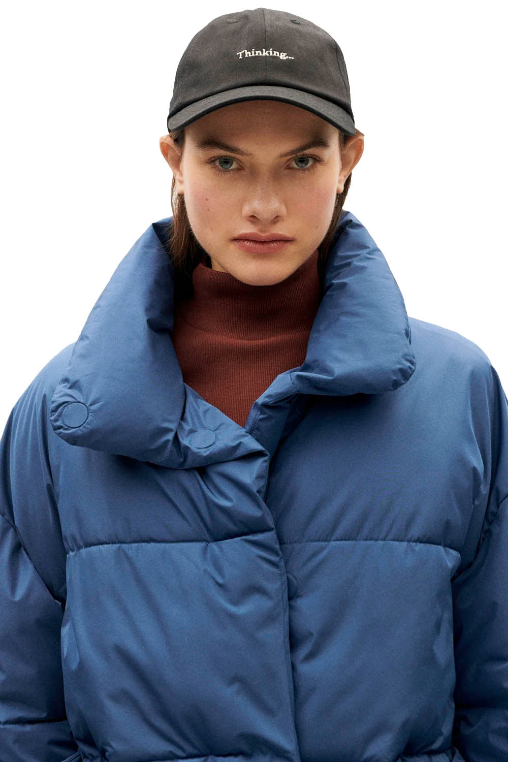 A model wearing a blue puffer jacket with a high padded collar, a brown turtleneck, and a black baseball cap with white embroidered text, set against a plain white background.