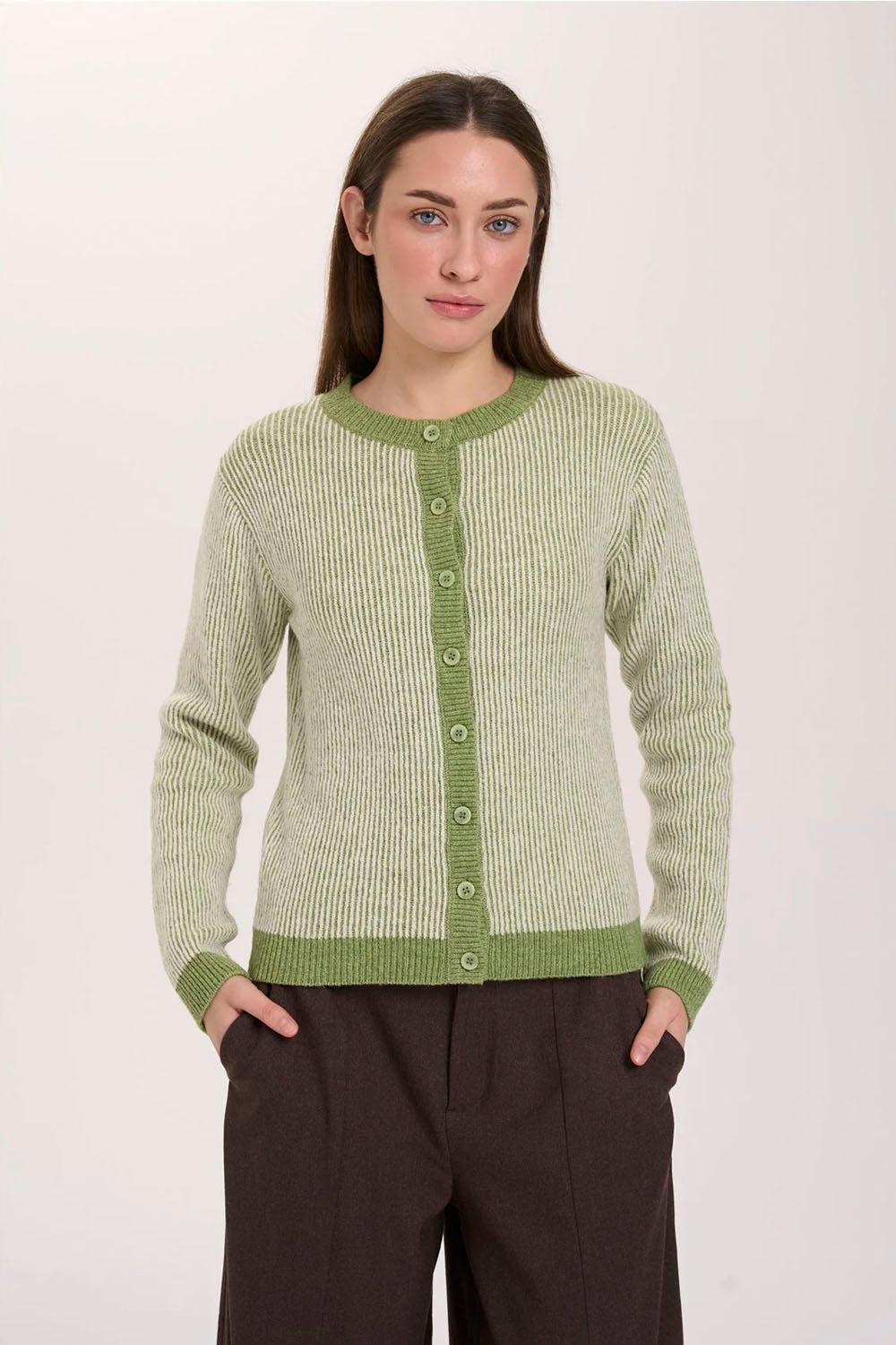A woman modeling a light sage green ribbed knit cardigan sweater with a darker green button placket, collar, cuffs, and hem, paired with dark brown trousers, against a plain off-white background.