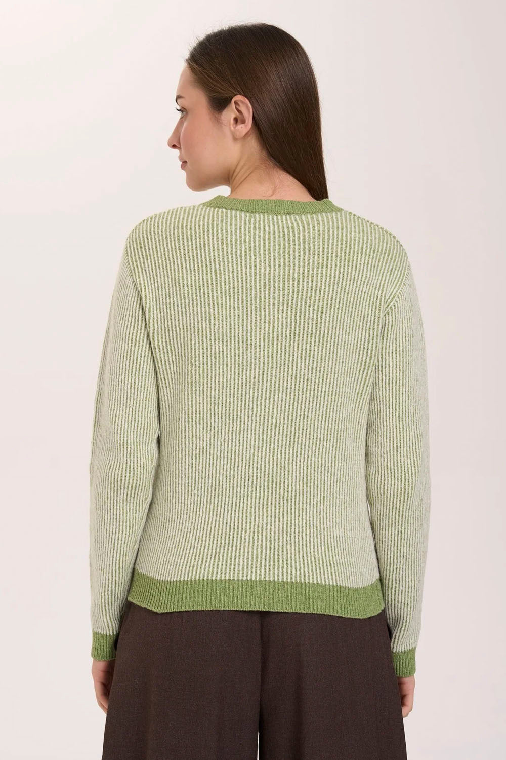 A woman shown from the back wearing a light sage green ribbed knit sweater with contrasting olive green trim at the neckline, cuffs, and hem, paired with dark brown trousers against a plain off-white background.