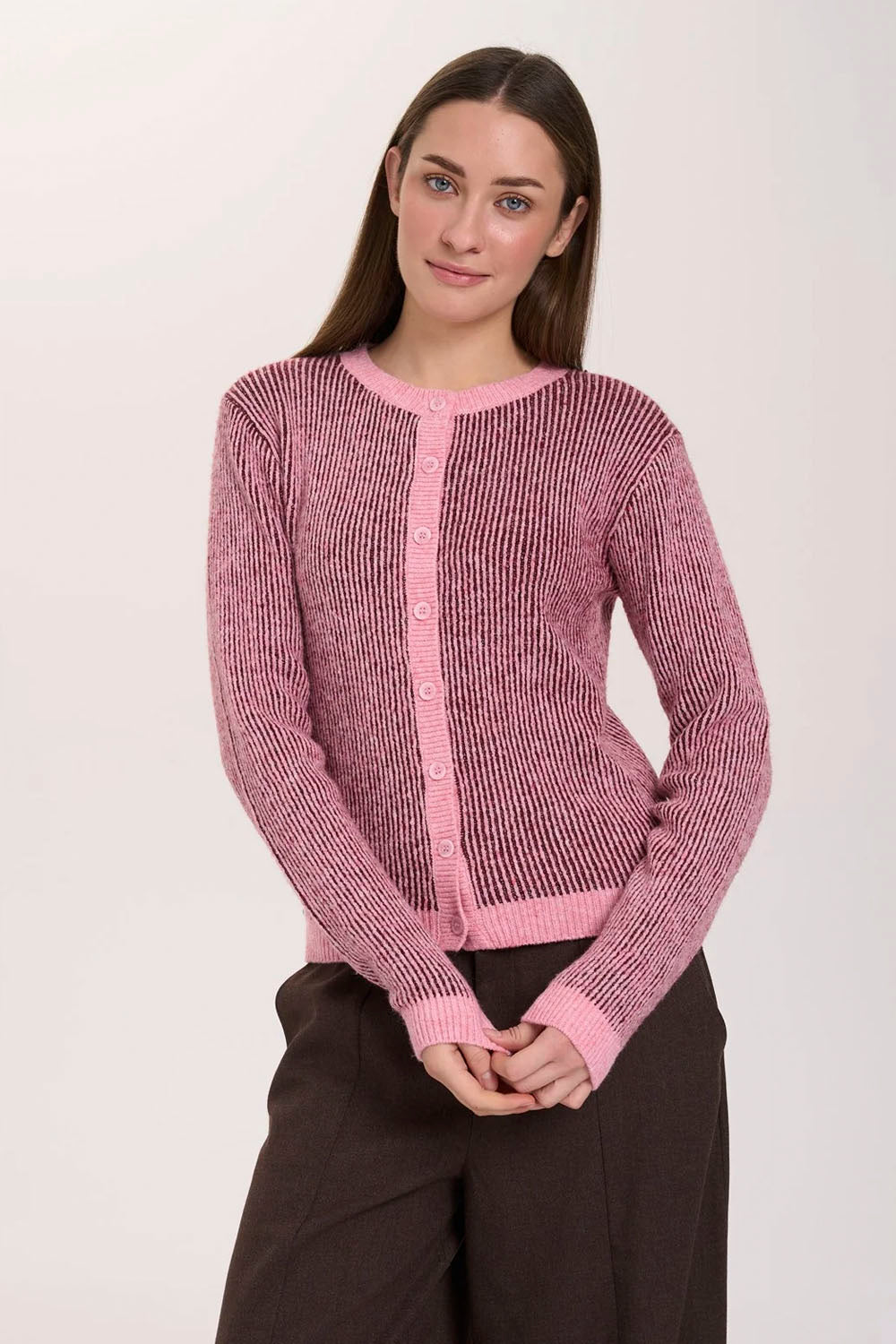 A woman modeling a pink ribbed knit cardigan sweater with a button-front closure and dark brown pants, photographed against a plain light background.