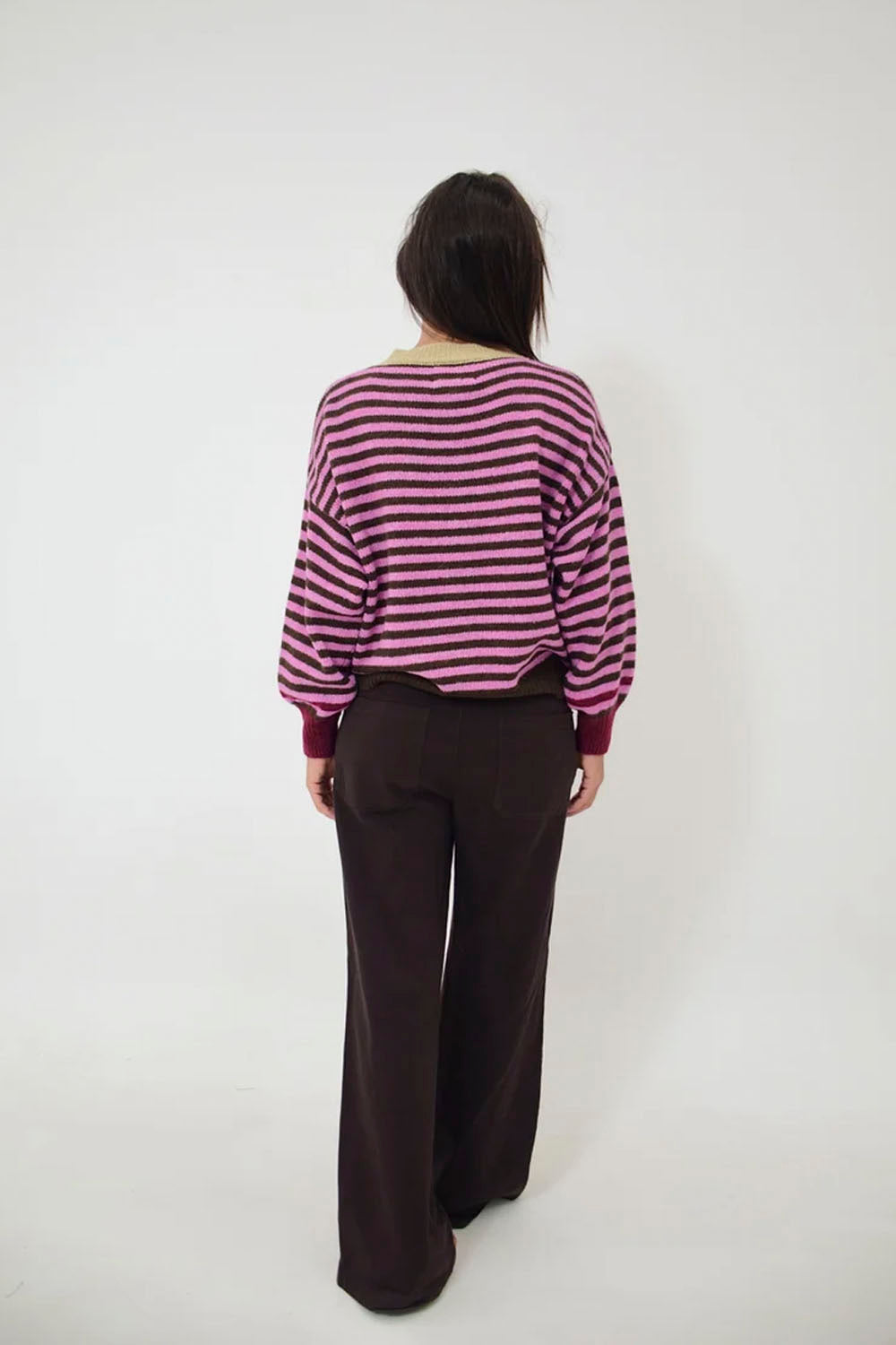 A person viewed from the back wearing a purple and pink striped long-sleeve top and dark brown wide-leg pants, standing against a plain white studio background.