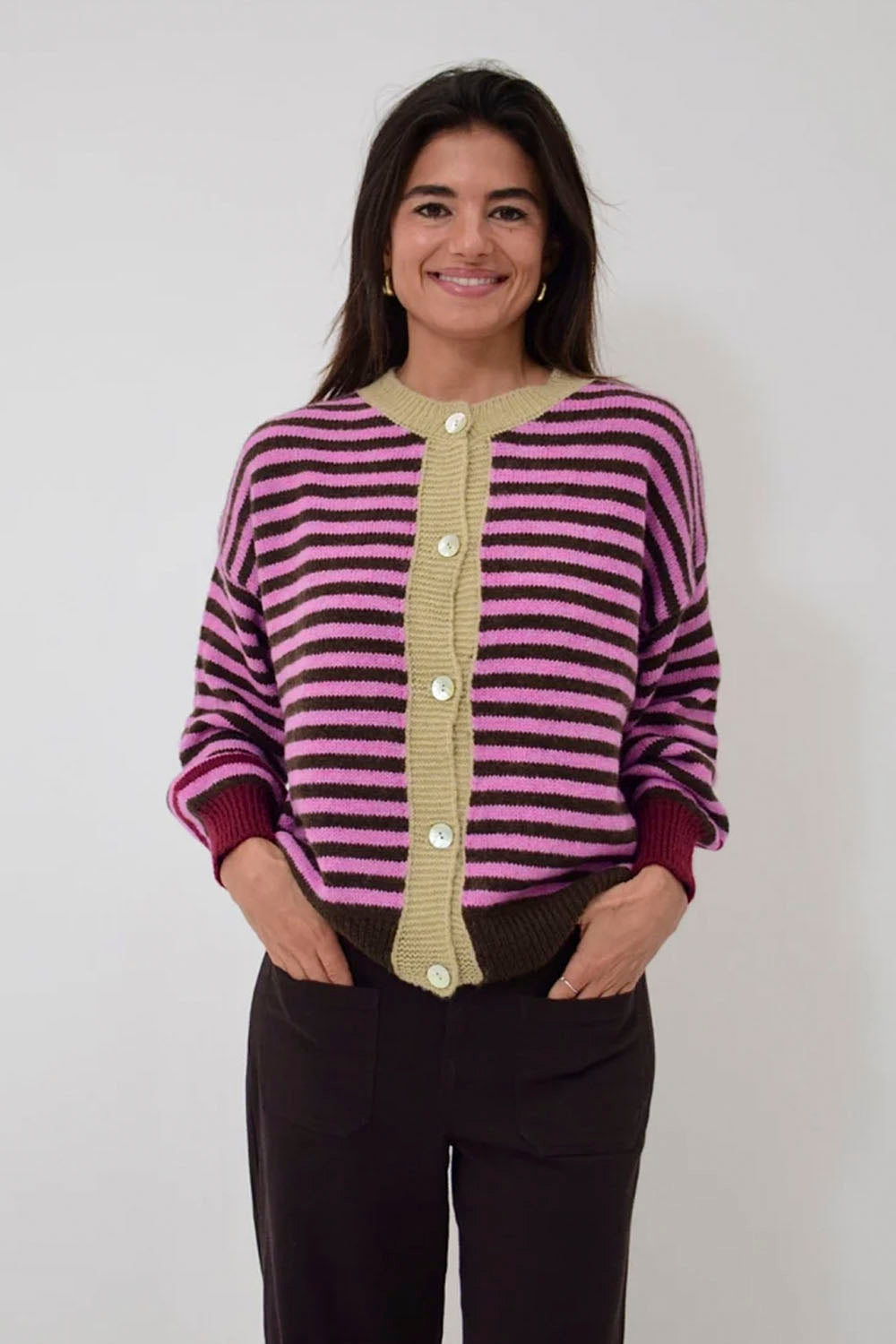 A woman wearing a pink and purple striped knit cardigan with a beige button placket, paired with dark brown pants, standing against a plain light gray background.
