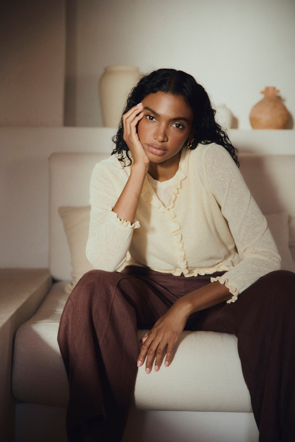 Woman sitting on a white couch wearing a yellow cardigan and brown pants.