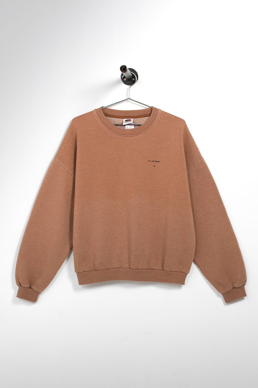 A brown crewneck sweatshirt hanging on a black hanger against a white wall. The sweatshirt has a small black embroidered logo on the left chest.