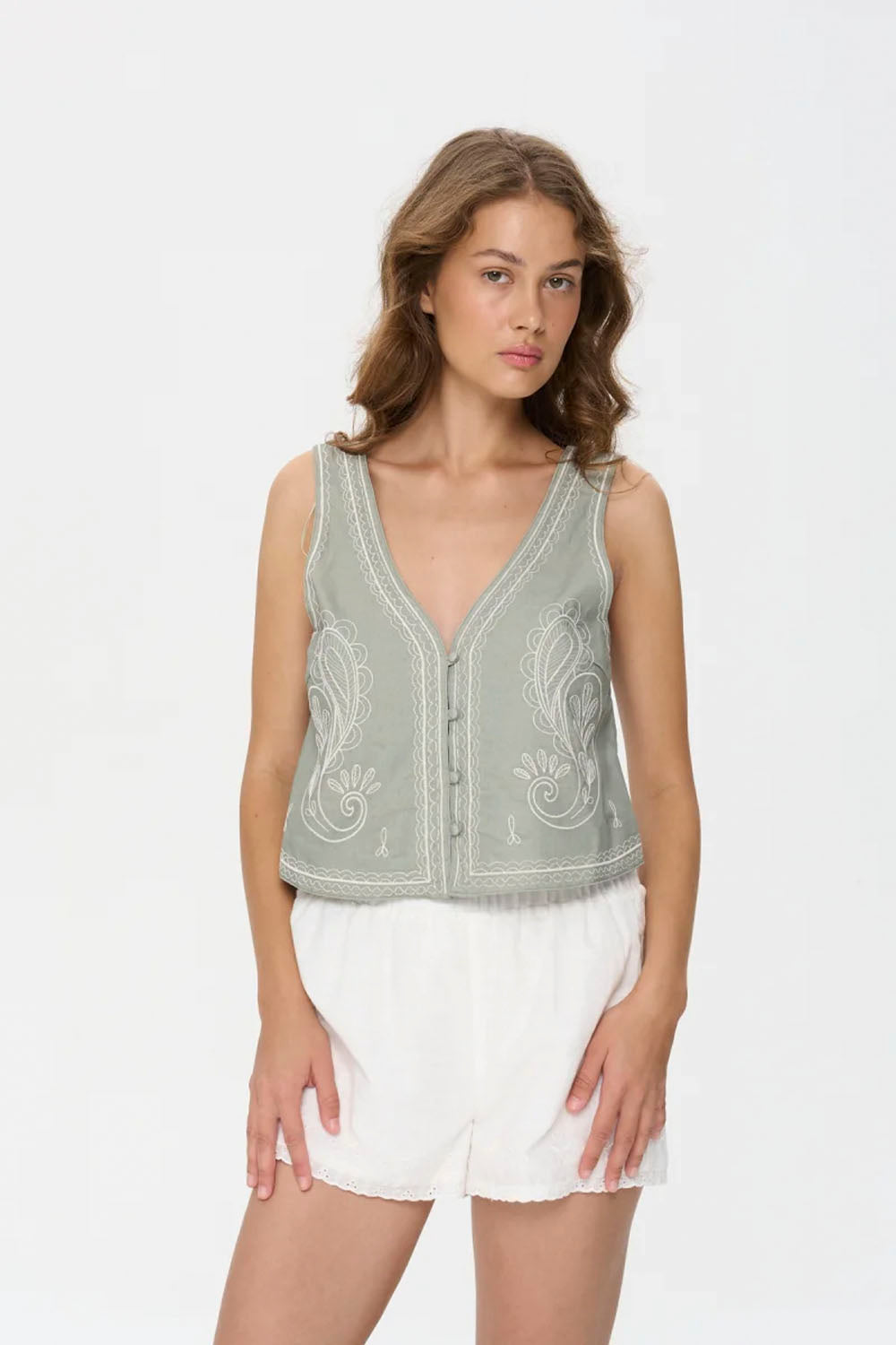Woman wearing a sleeveless embroidered top and white shorts on a plain background