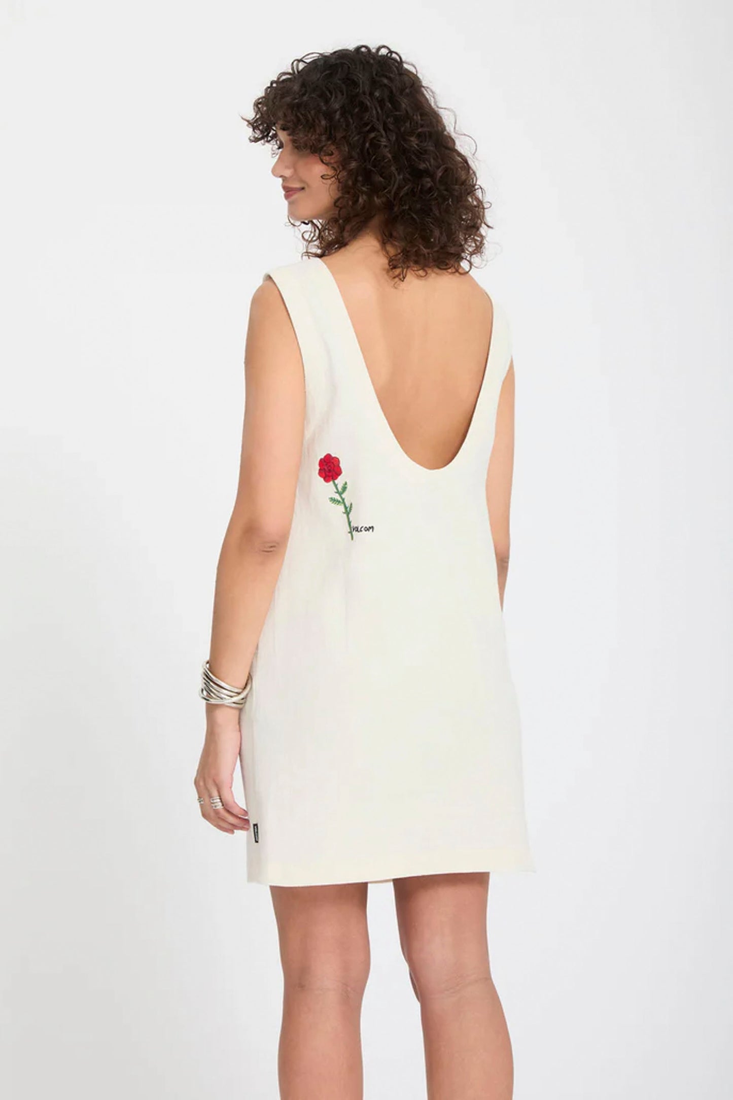 Woman wearing a white Volcom Fa Mickey Mason dress with a red rose embroidery on a plain background