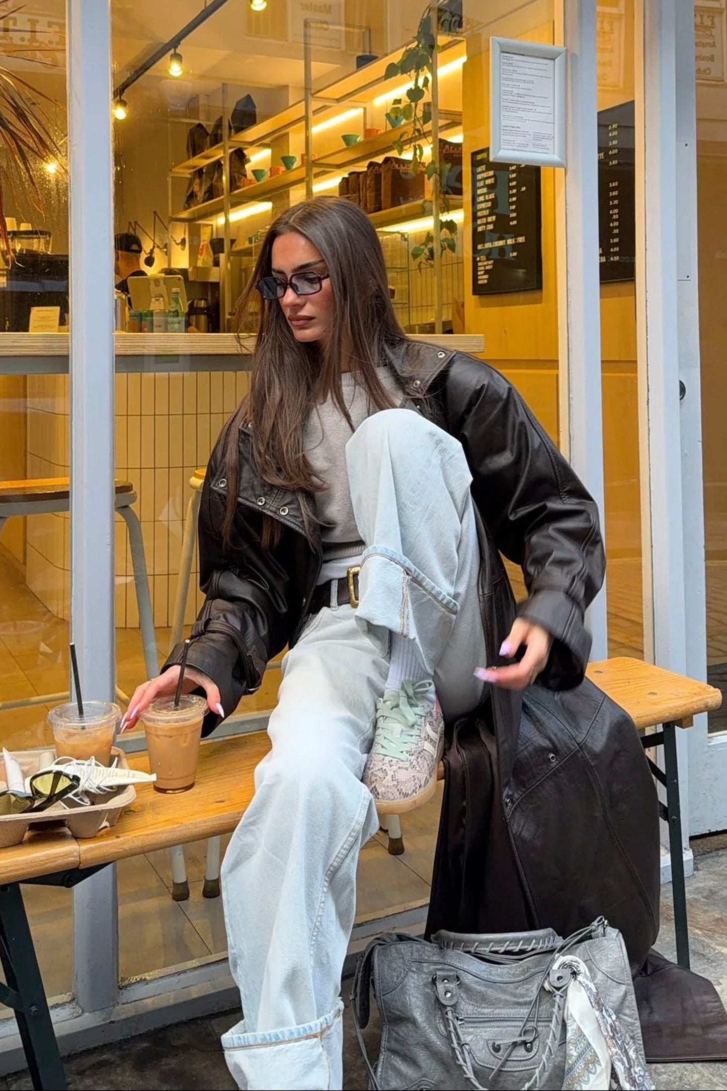 Woman sitting at a table in a cafe wearing a black coat, white pants and Coolway Goal Roar Flux sneakers