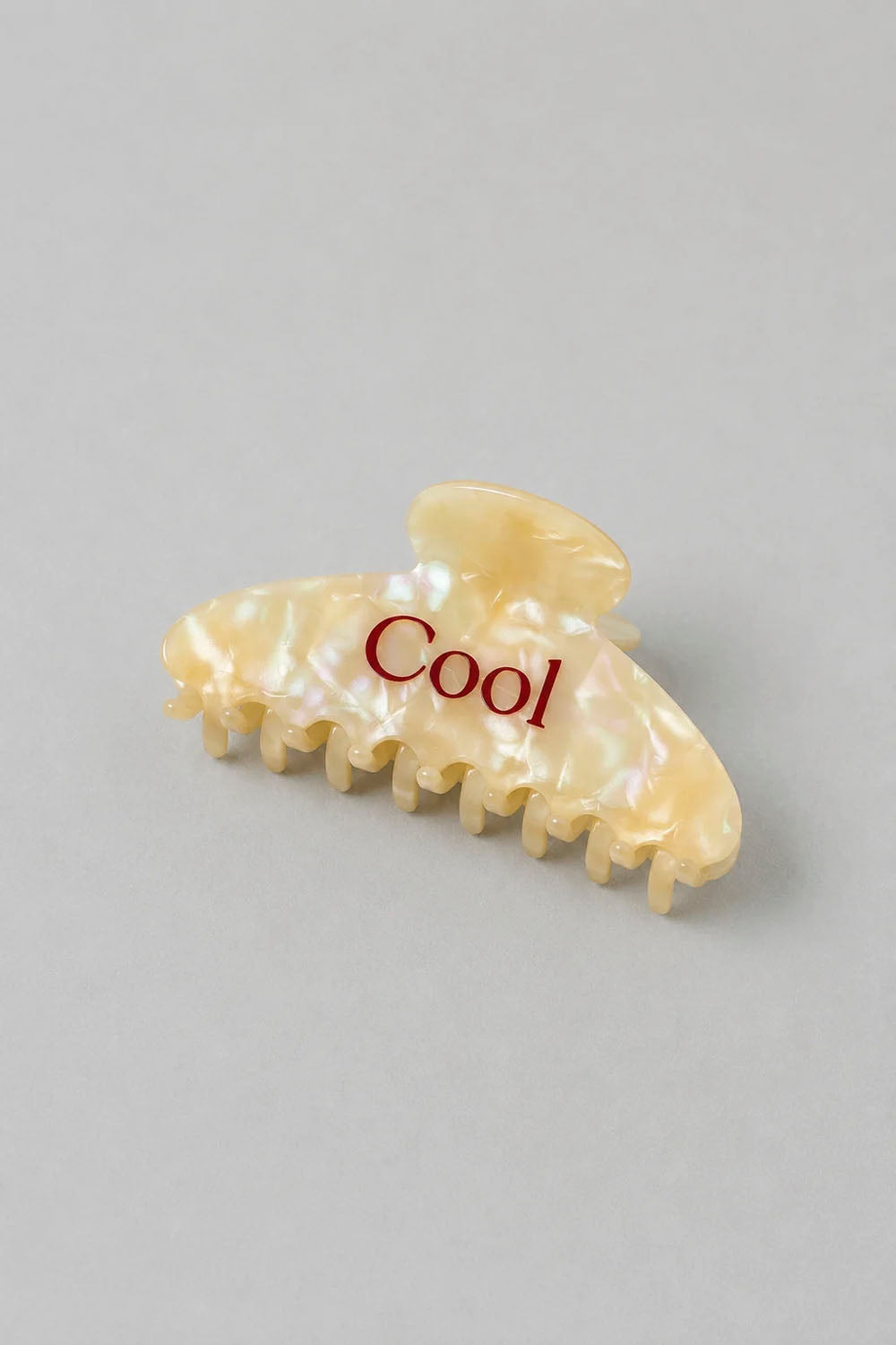 A single hair claw clip in a pearlescent cream shell-like finish with the word 'Cool' printed in red across the front, photographed on a plain light gray background.
