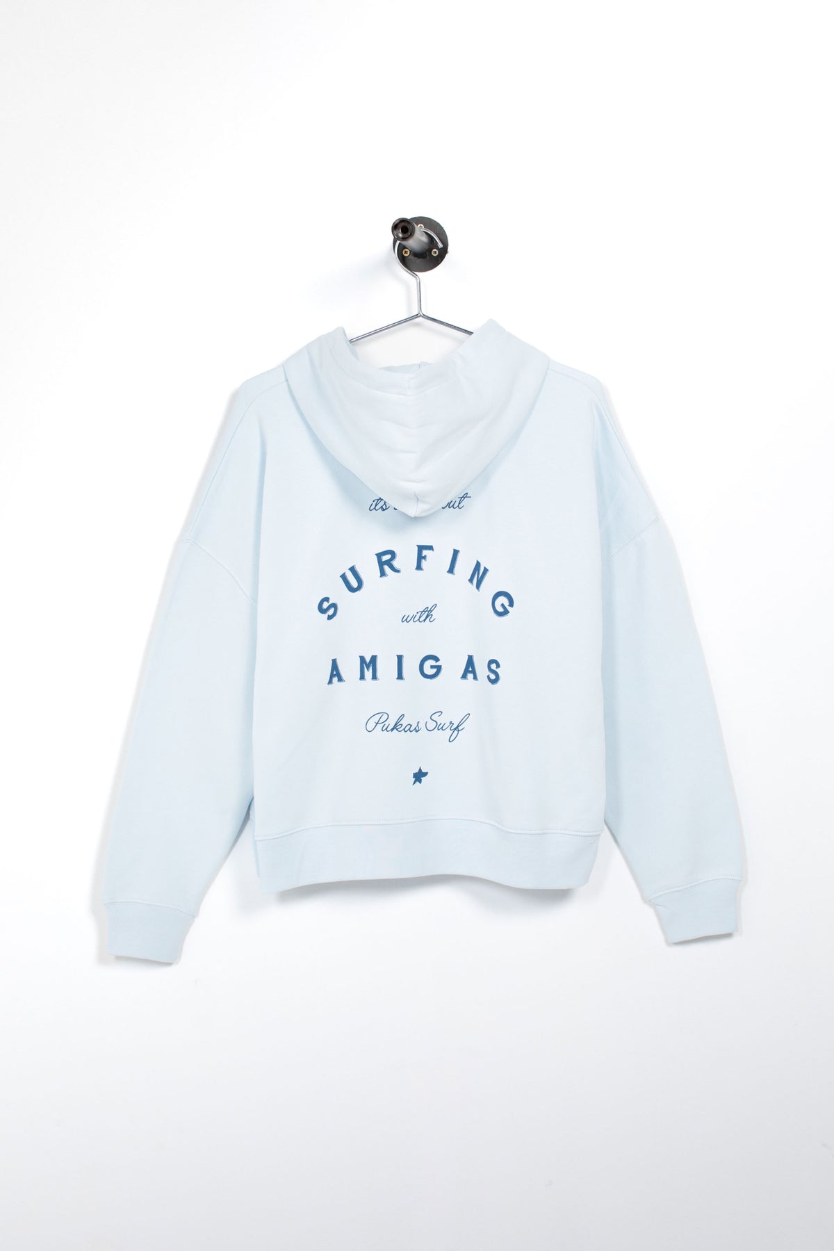 A light blue cropped hoodie hanging on a white hanger against a plain white wall. The back of the hoodie features blue text that reads 'SURFING with AMIGAS' and 'Palmie Surf' with a small star graphic below.