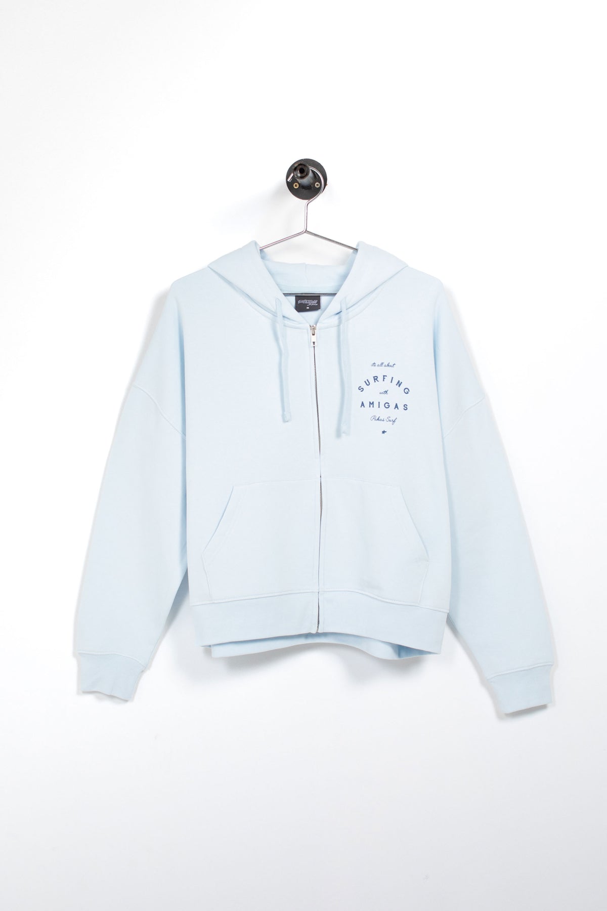 A light blue zip-up hoodie with a drawstring hood and kangaroo pockets, hanging on a metal hanger against a plain white wall. The left chest features a small dark blue graphic print.