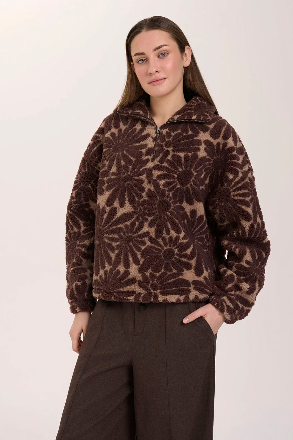 A woman modeling a brown and beige floral patterned fleece pullover with a half-zip collar, paired with dark brown wide-leg trousers, against a plain light background.