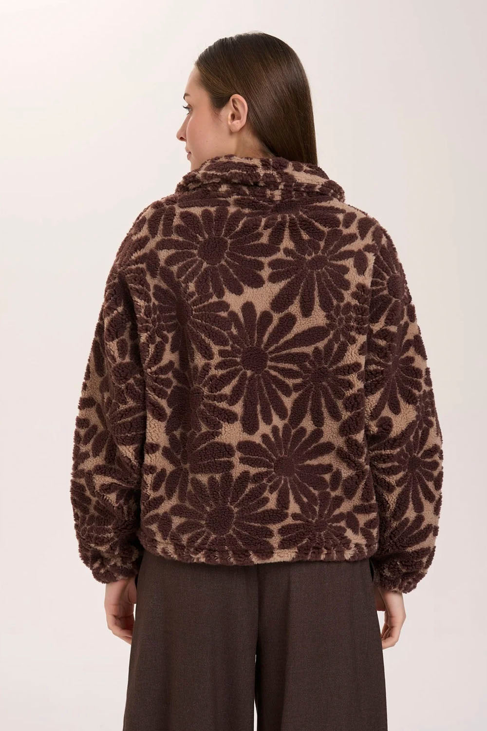 A woman modeling a brown and beige floral patterned fleece pullover with a half-zip collar, paired with dark brown wide-leg trousers, against a plain light background.