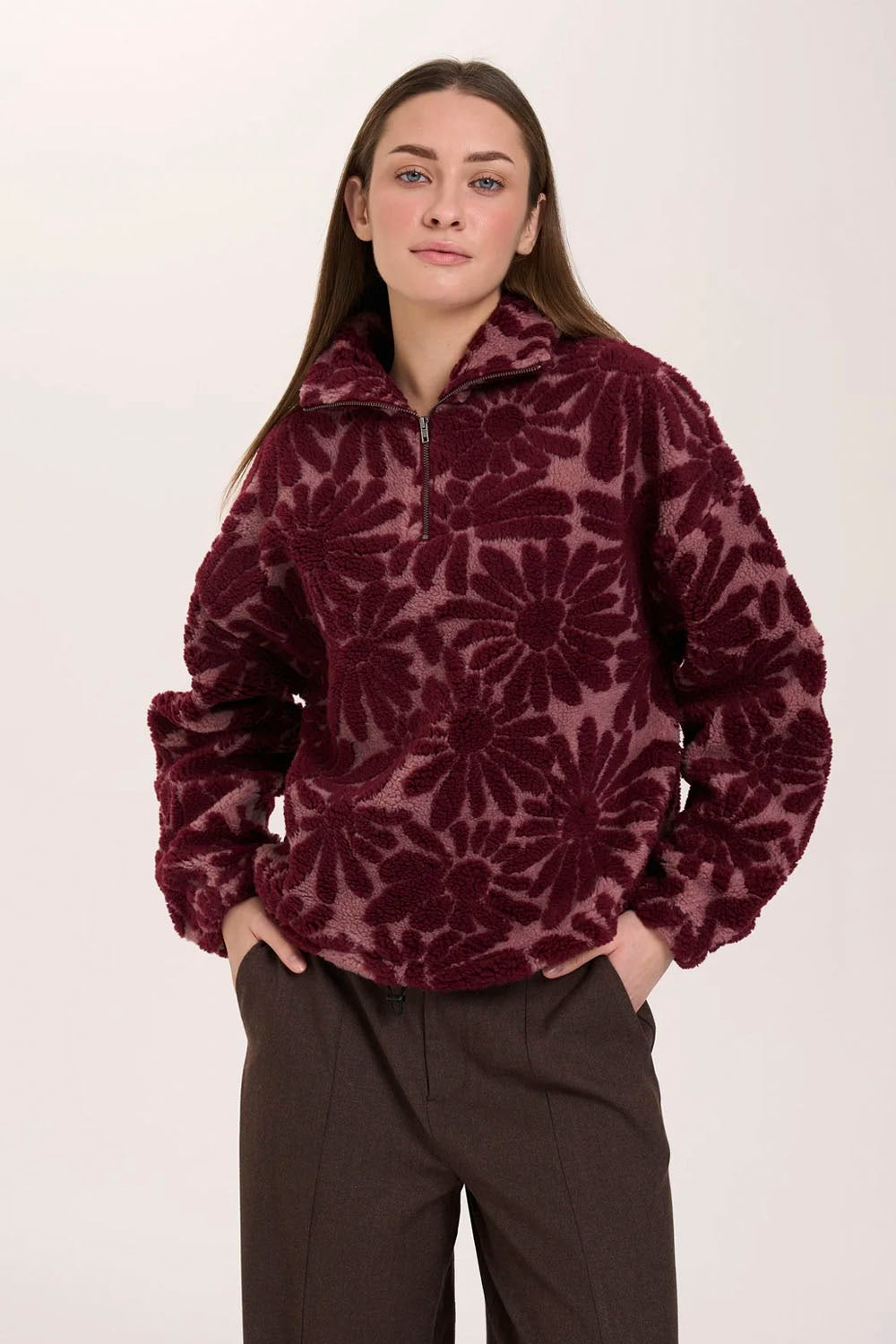 A woman modeling a dark burgundy long-sleeve quarter-zip fleece pullover with a tonal floral pattern, paired with dark brown trousers, against a plain light background.