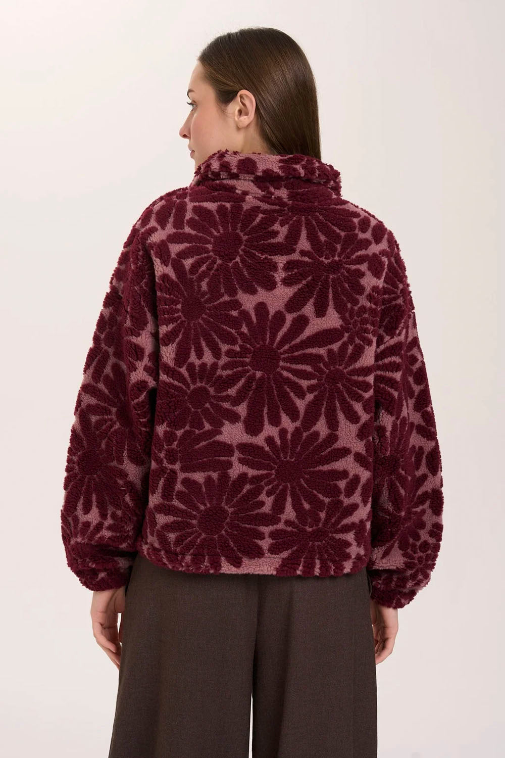 A woman shown from the back wearing a fuzzy cropped jacket with a large floral pattern in deep burgundy and plum, paired with dark wide-leg pants, against a plain light background.