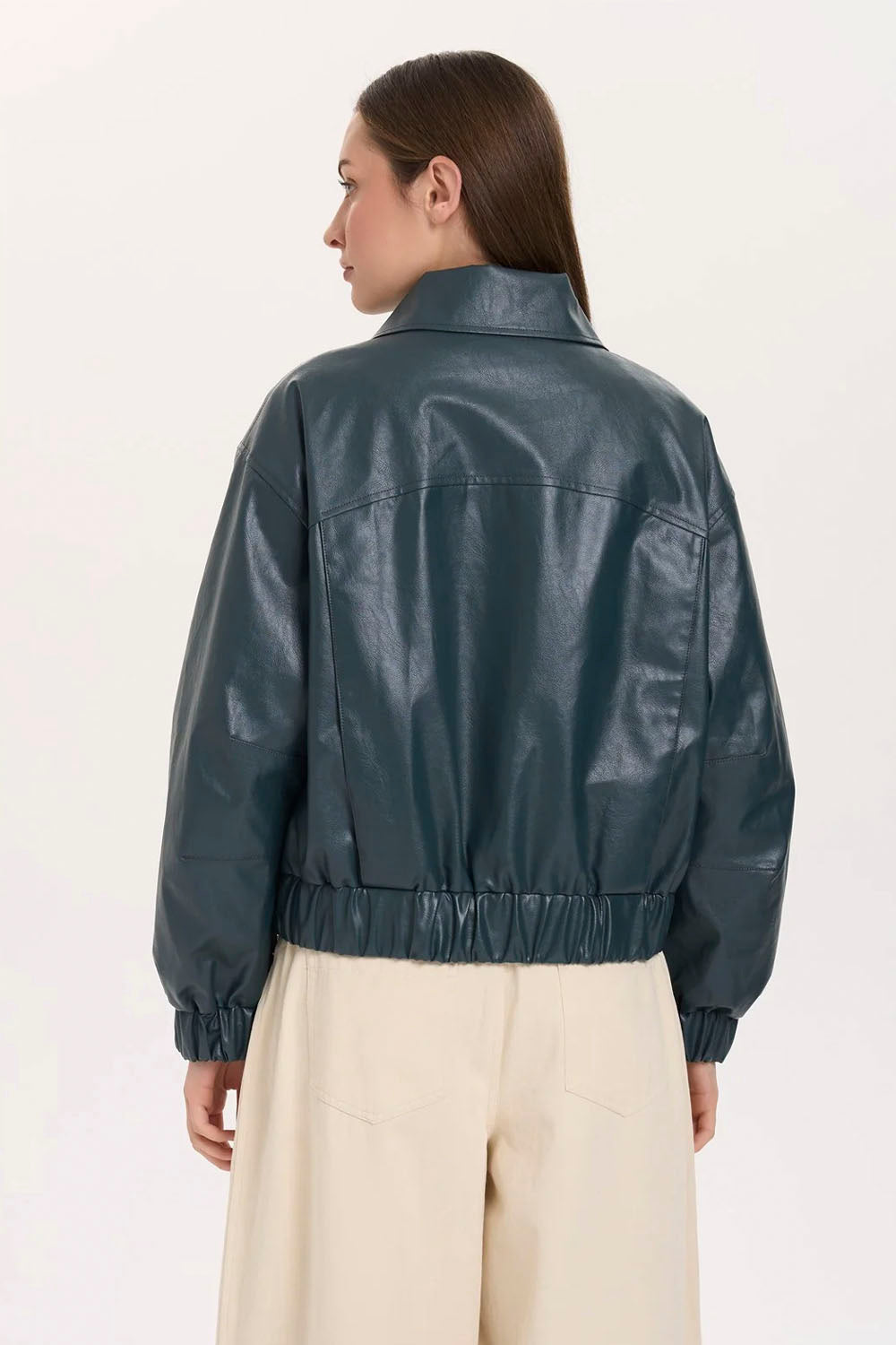 A woman shown from the back wearing a dark teal faux-leather jacket with a gathered elastic hem and cuffs, paired with cream wide-leg trousers, against a plain off-white studio background.