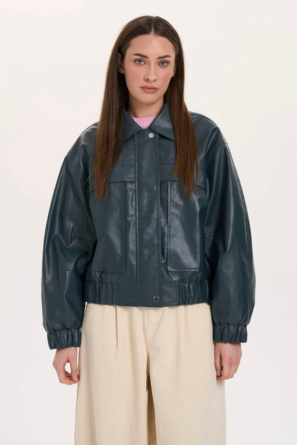 A woman modeling a dark teal faux-leather jacket with a button-up front, chest pockets, and elasticized cuffs, paired with cream wide-leg trousers against a plain white studio background.