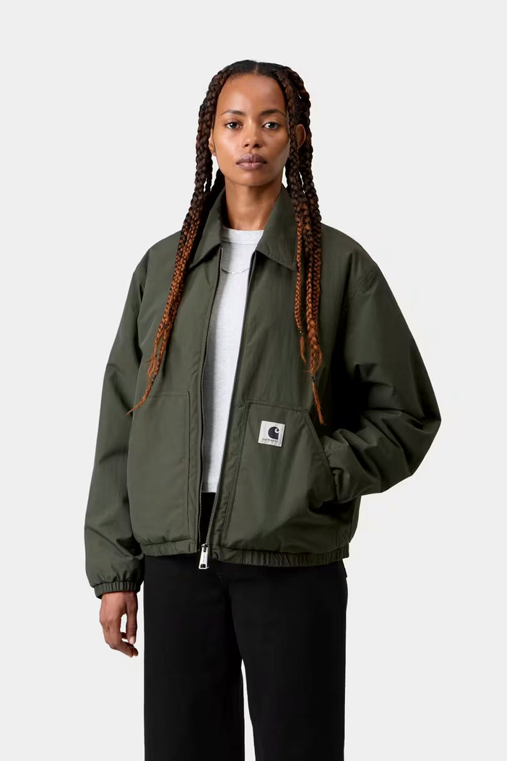 A model with long braided hair wearing an olive green Carhartt jacket over a white t-shirt and black pants, standing against a light gray studio background.