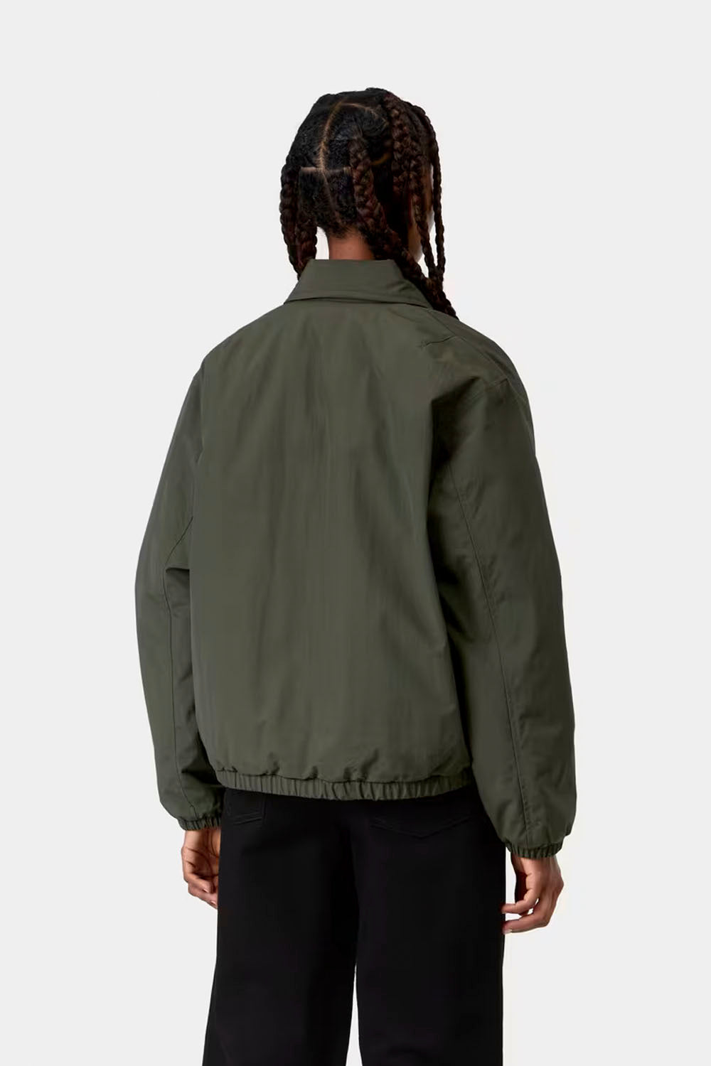 A person shown from the back wearing an olive green jacket and black pants against a light gray studio background.