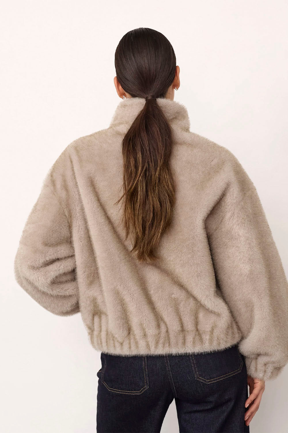 A woman shown from behind wearing a fluffy, oversized beige faux-fur jacket and dark blue jeans against a plain white background.